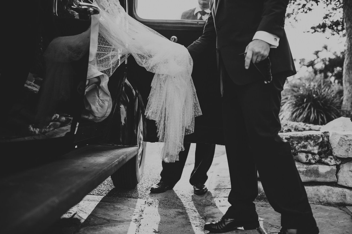 Black and white photo of bride in wedding dress stepping out of vintage car assisted by groom.