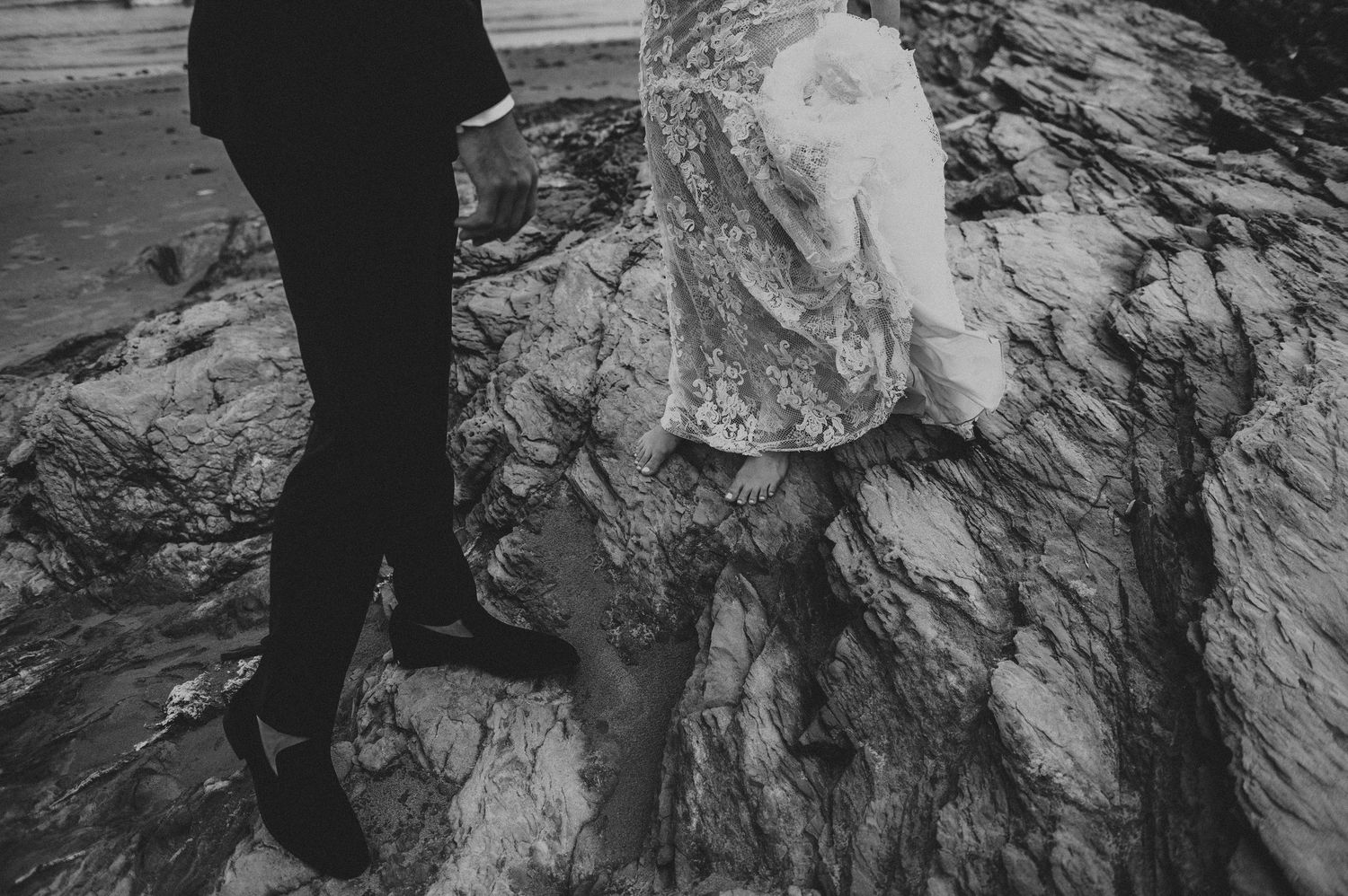 Black and white close-up of bride in lace gown and groom navigating rocky terrain.