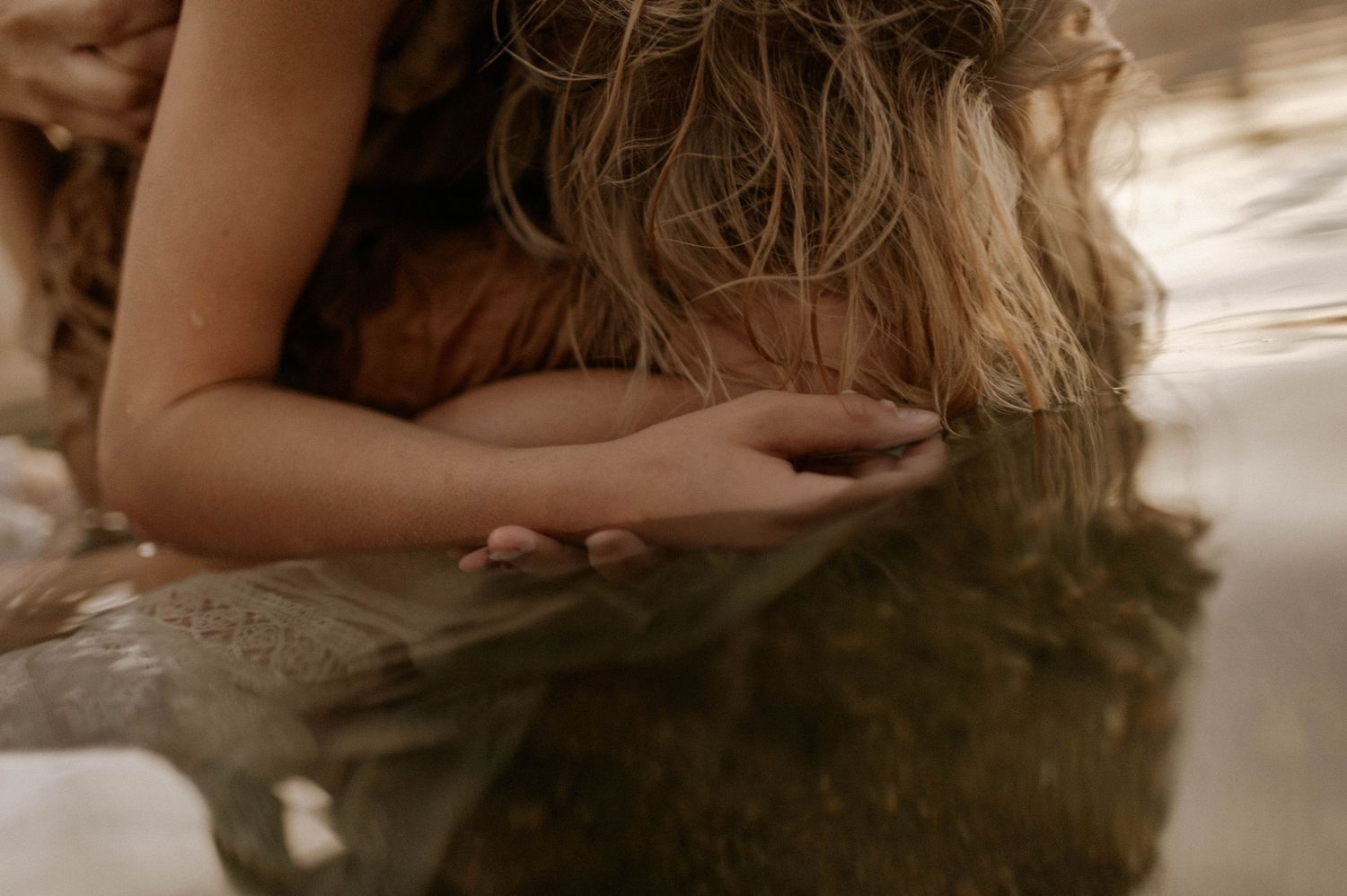 Close-up of a persons hands gently touching calm water while wearing a brown flowing dress.