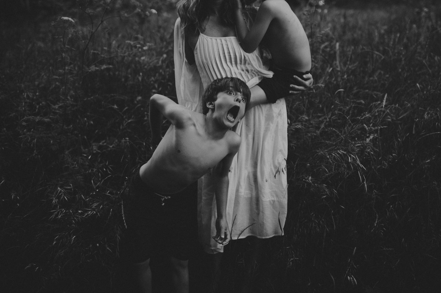 Black and white photo of a playful shirtless boy with open arms standing beside a woman in a white dress.