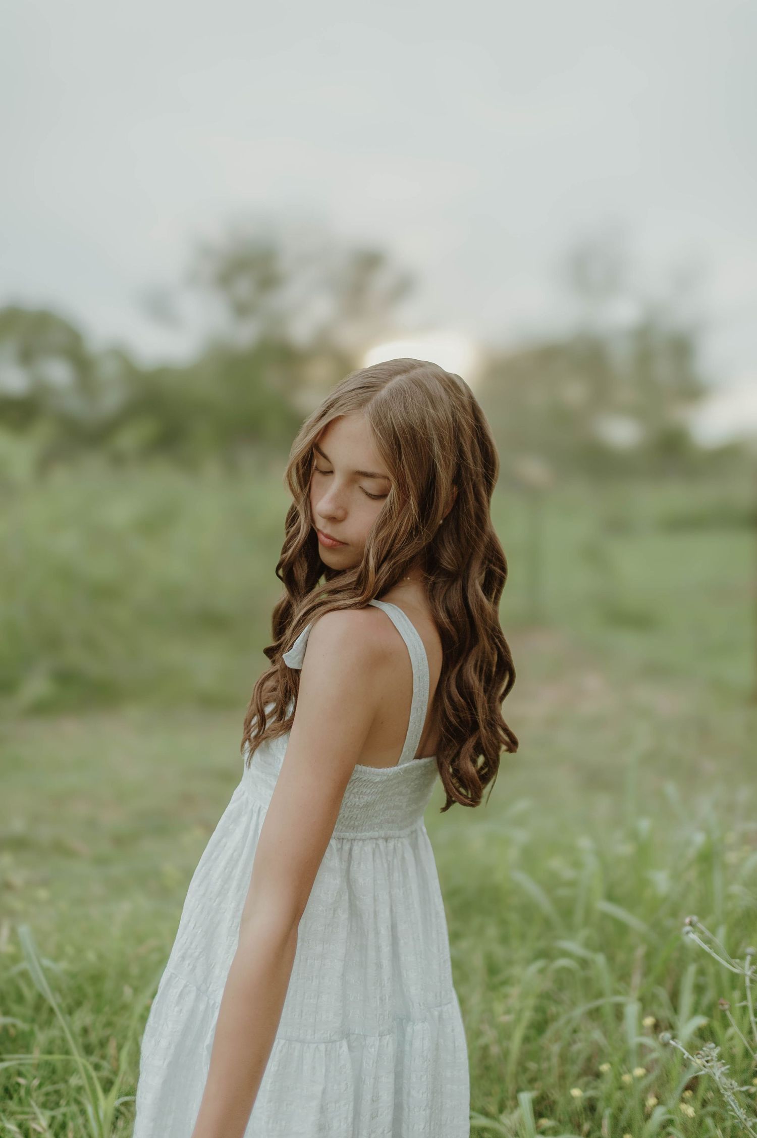 Young girl with long wavy brown hair in a white sundress standing in a green meadow at sunset.