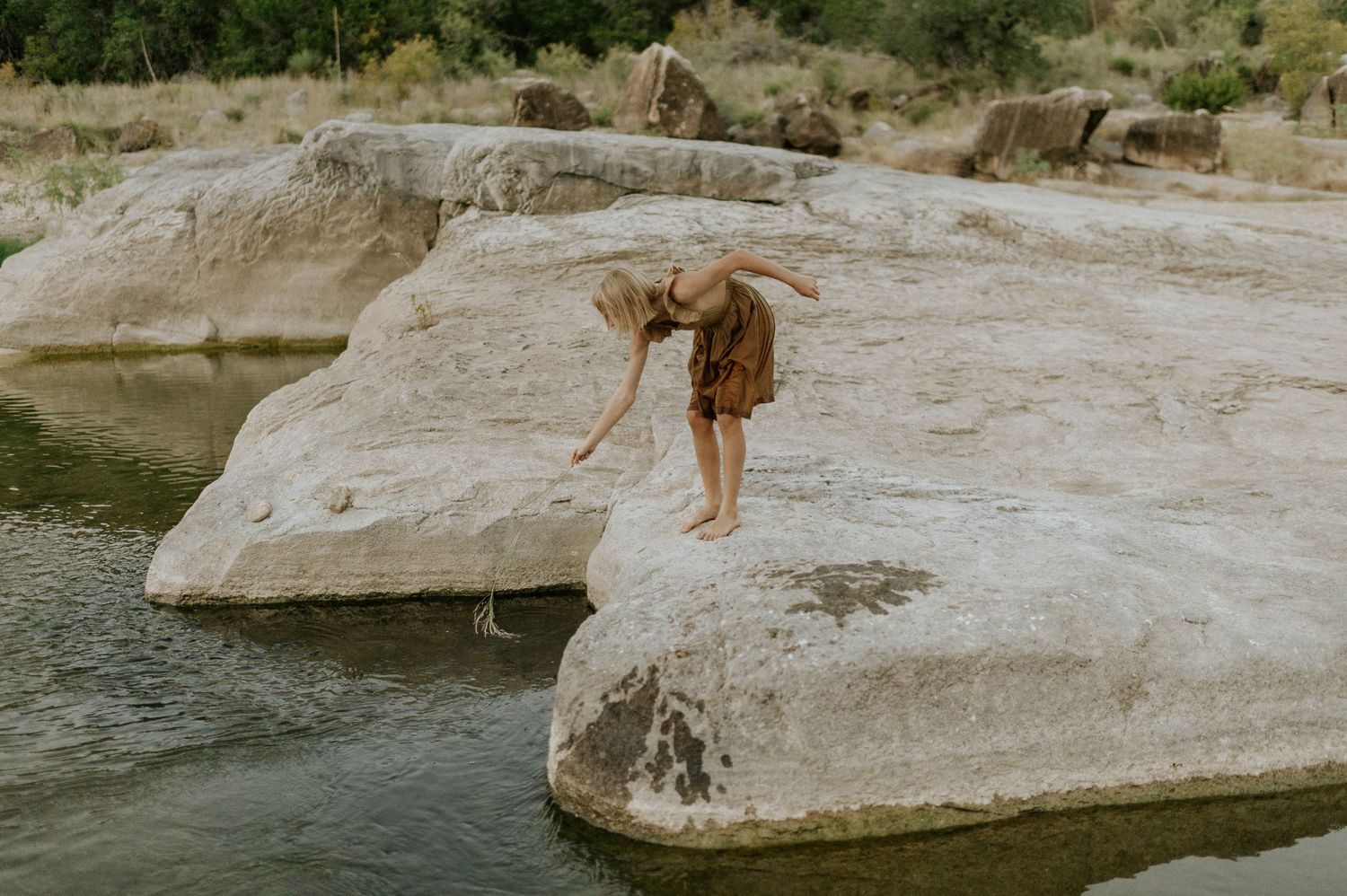 Child in brown dress leaning over rock edge to touch water at a scenic river location.