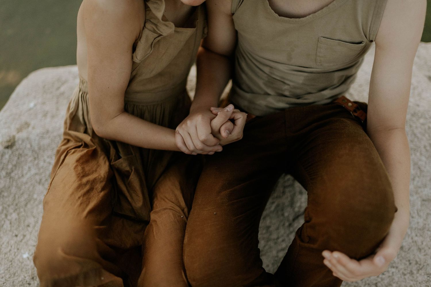 Two people sit together on a rock, holding hands in warm earth-toned clothing.