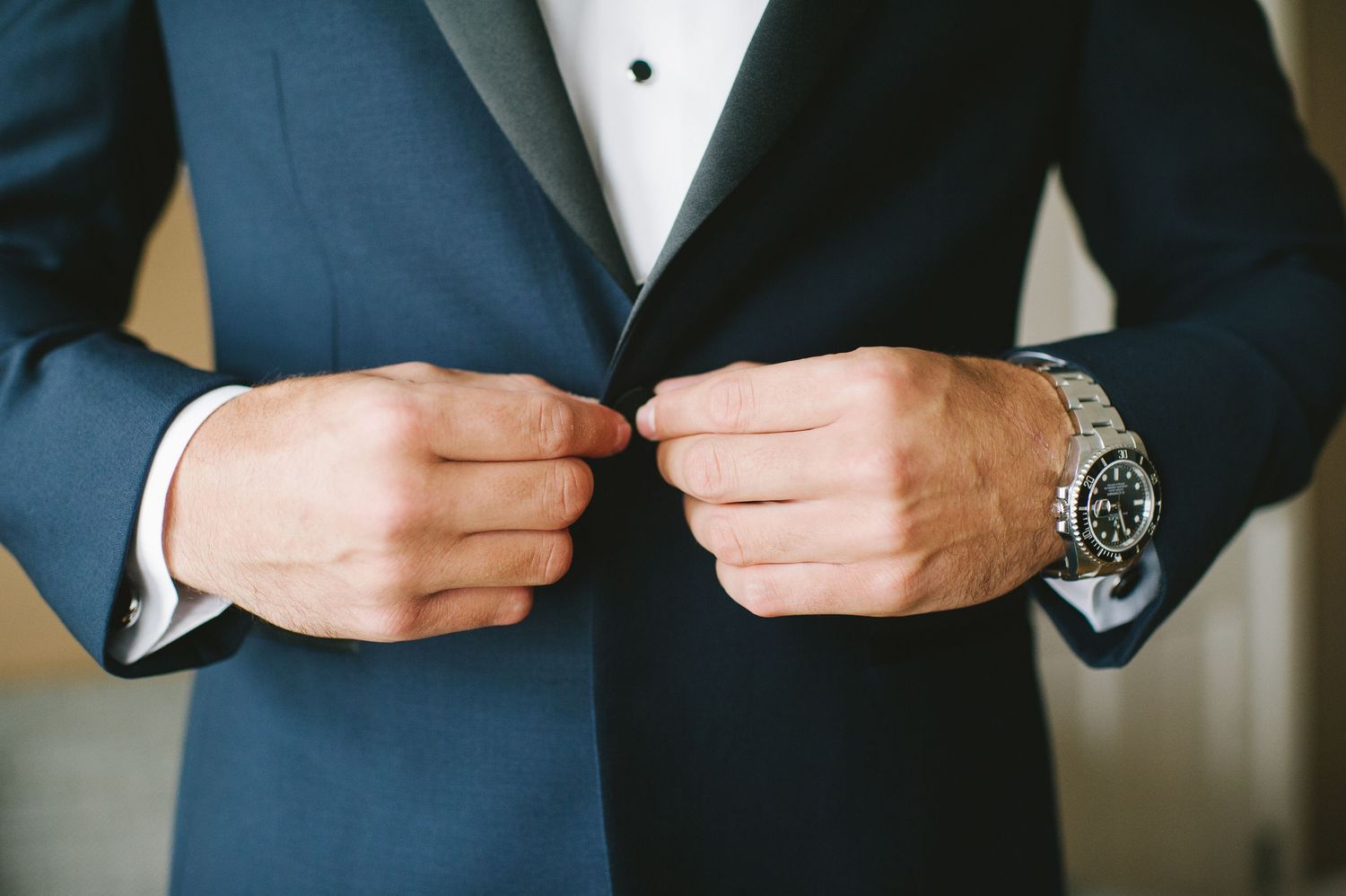 Groom in navy tuxedo adjusts jacket buttons showing elegant silver watch on wrist.
