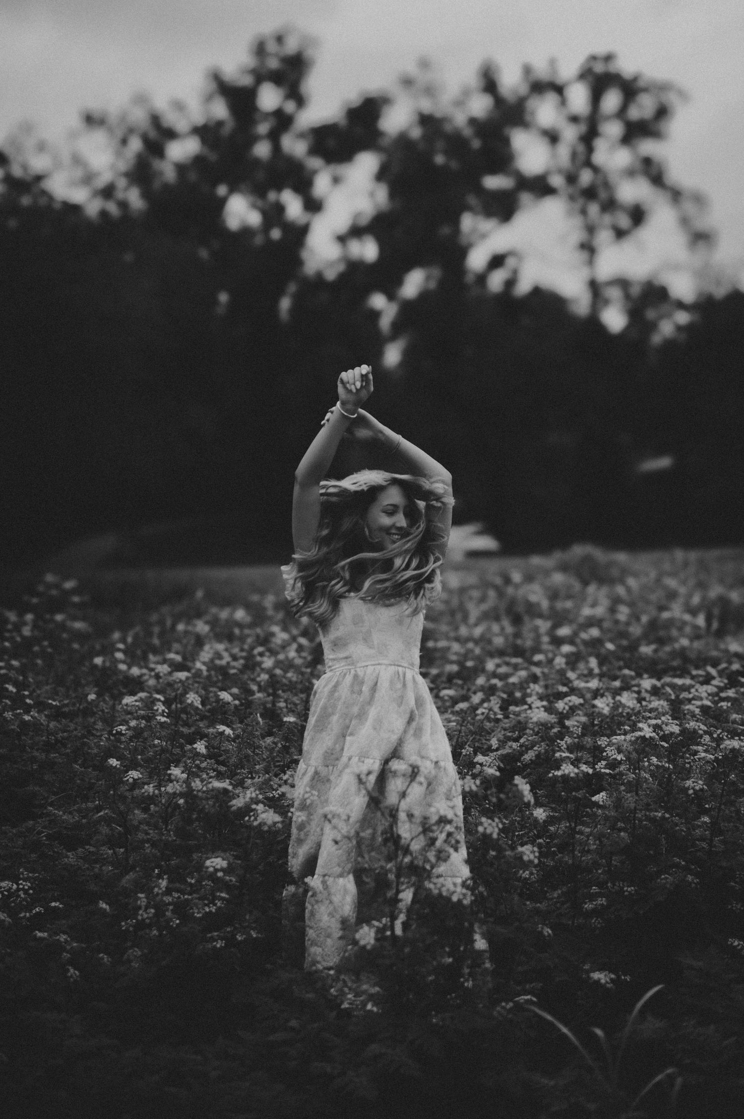 Black and white photo of a woman in a floral dress dancing joyfully in a wildflower field with trees in the background.
