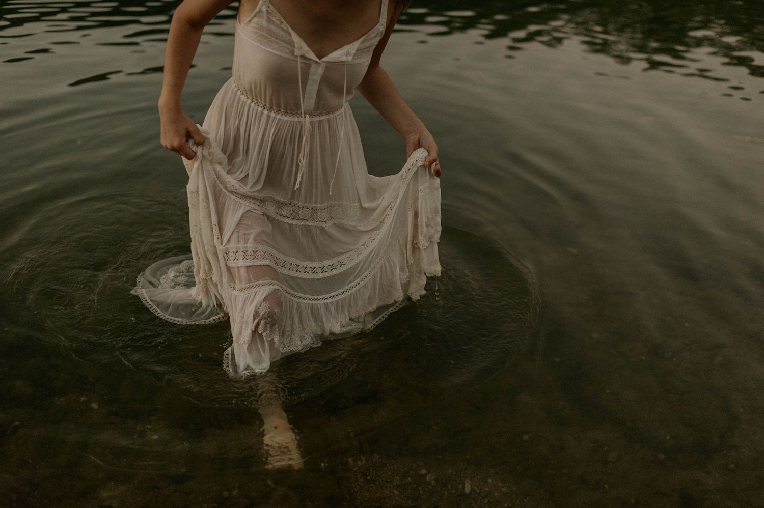 Woman in flowing white lace dress wading through calm dark water.