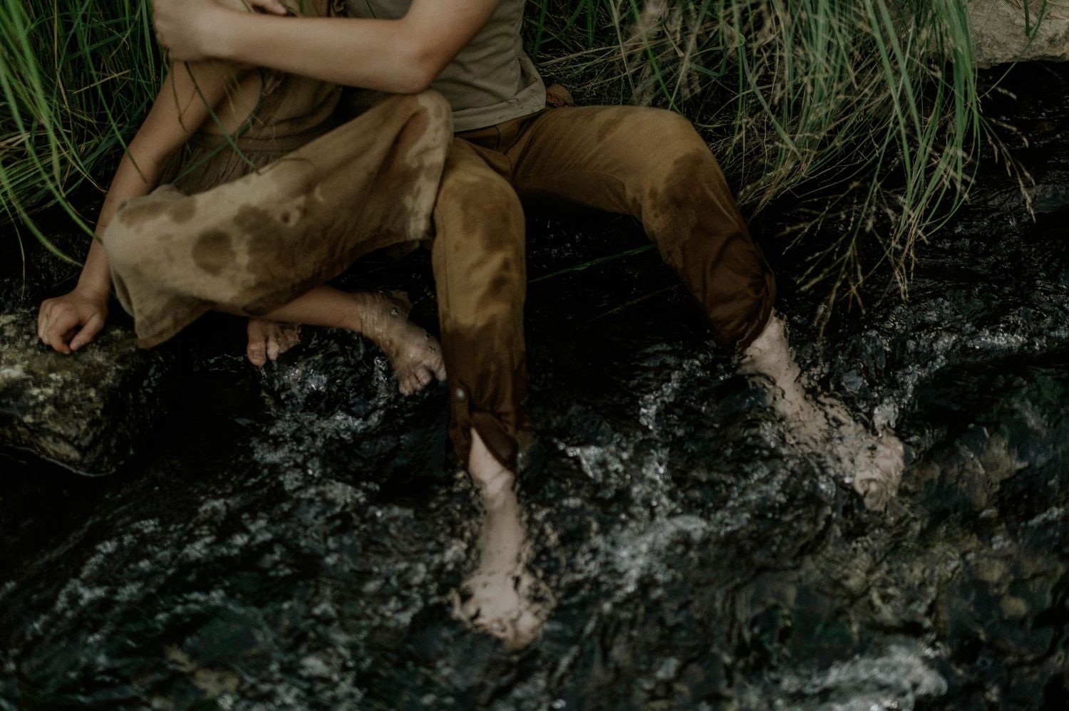 Two children sit barefoot on rocks splashing feet in a flowing stream surrounded by lush green grass.