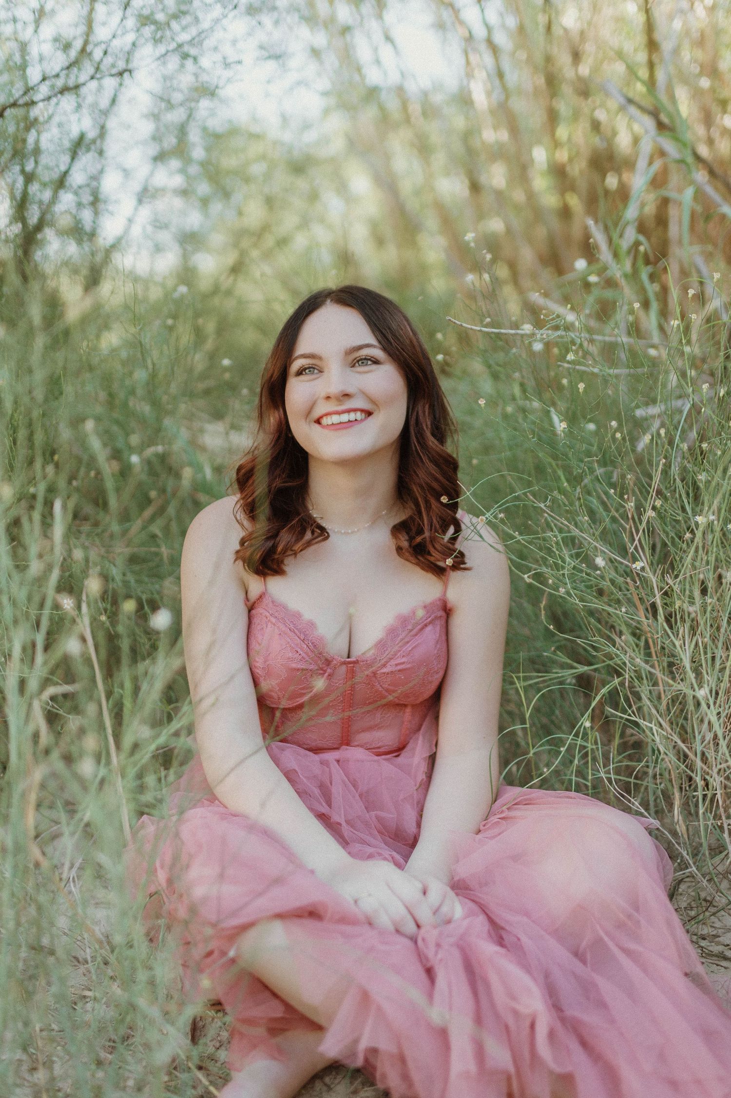 Smiling young woman in pink dress sitting outdoors among tall grasses in natural light.
