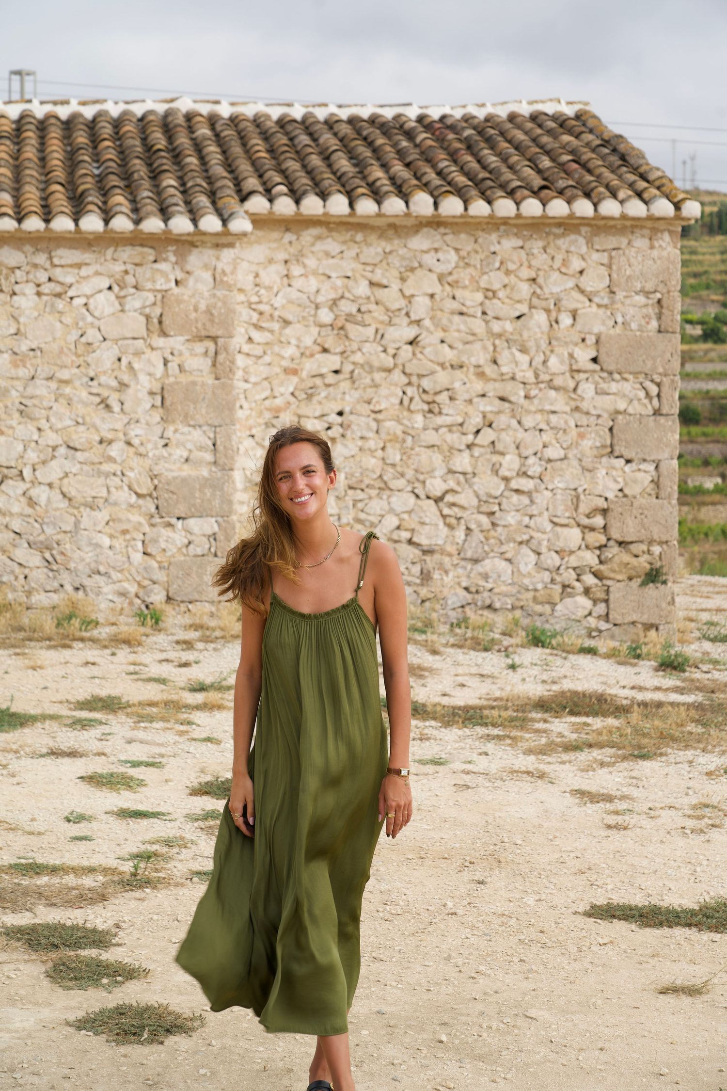 Vrouw in groene zomerjurk loopt voor rustiek stenen gebouw, fotografie door Eva Berendse