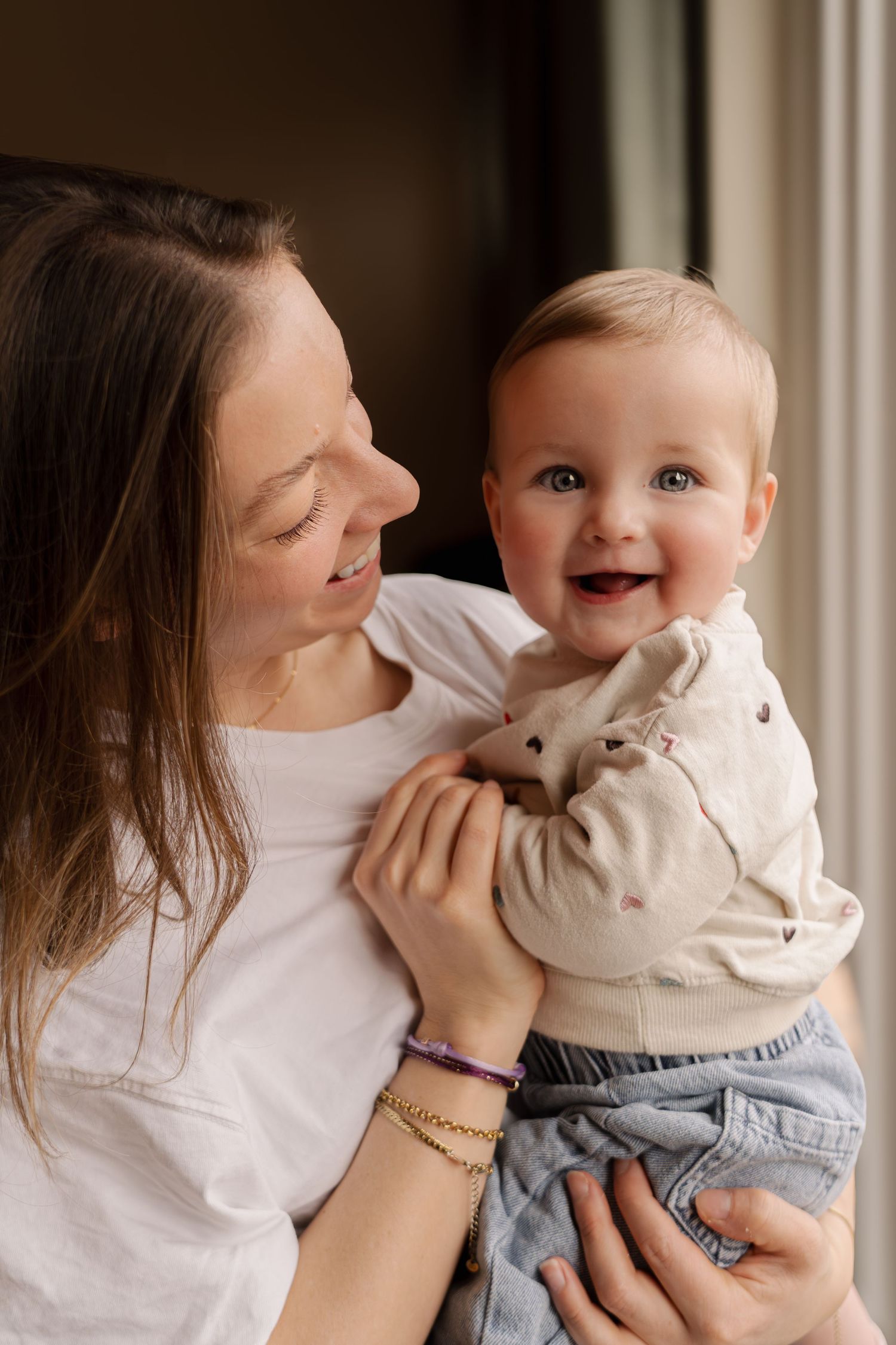 Moeder houdt lachende baby vast bij raam, newborn fotoshoot Milly en Lilly door Eva Berendse