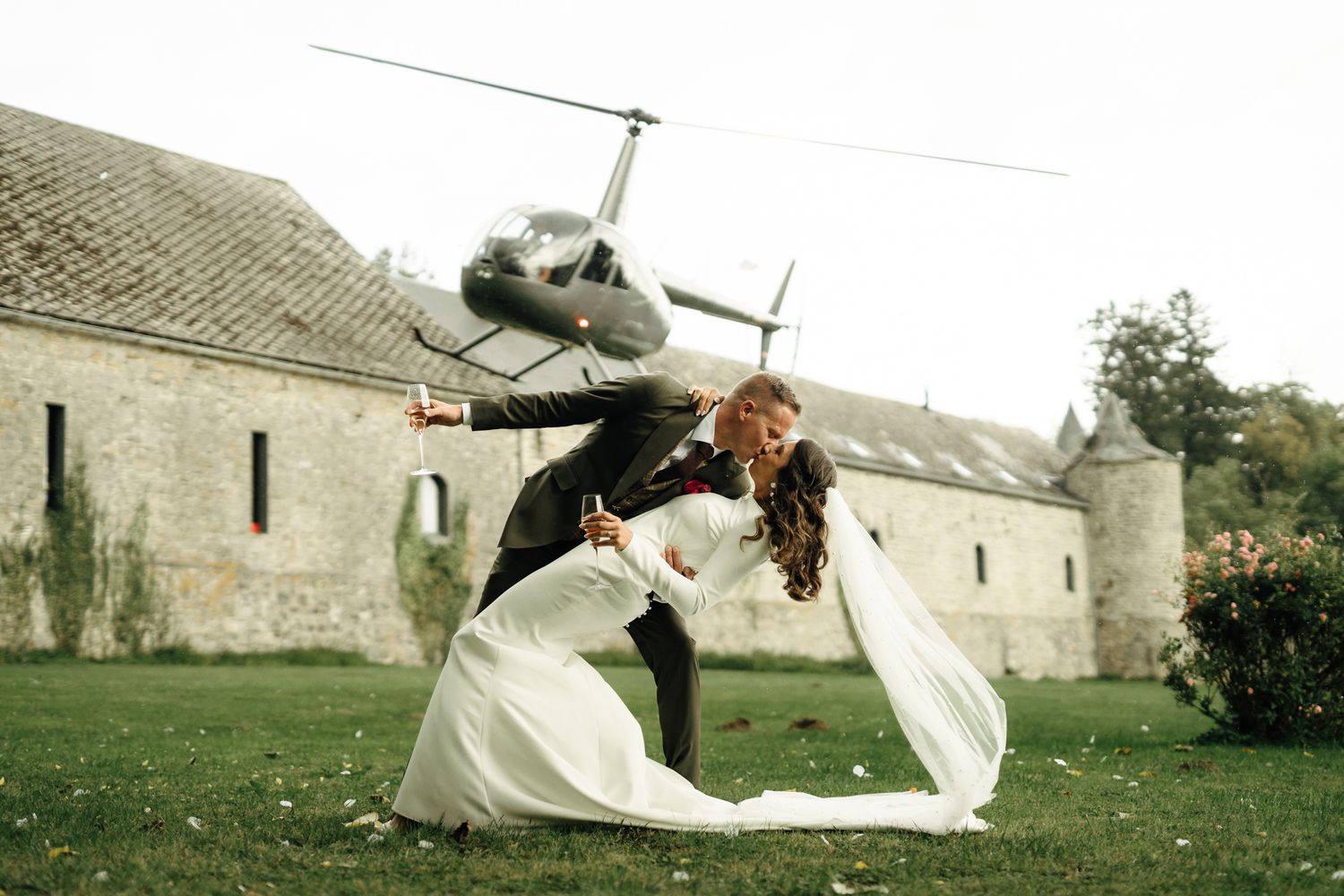 Bride and groom share a romantic dip kiss on lawn with helicopter and historic stone building backdrop.