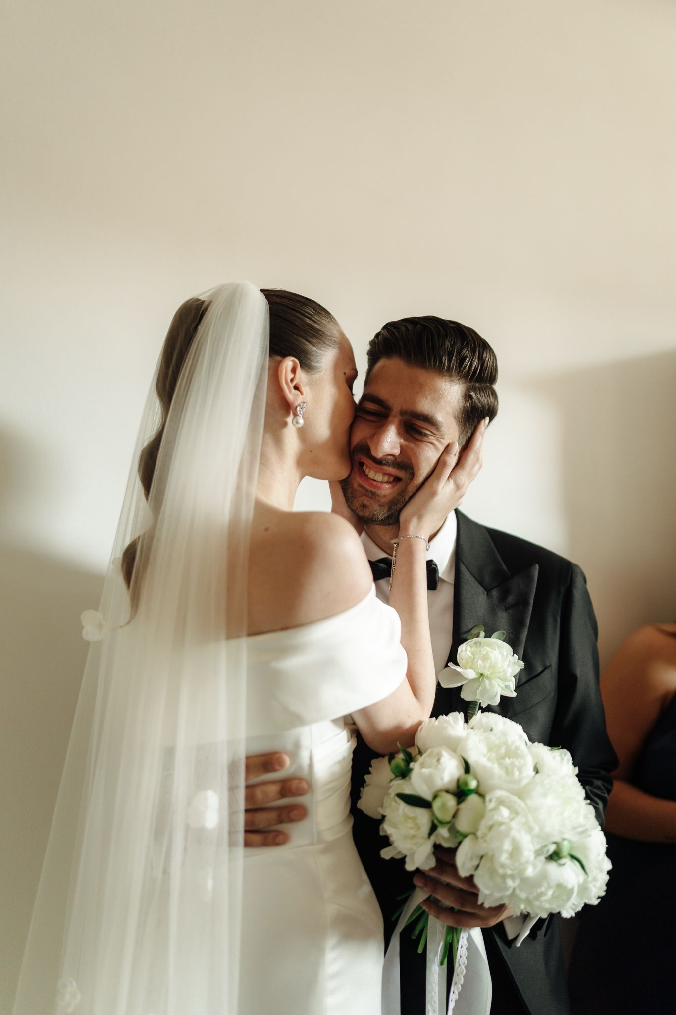 Smiling groom holds white peony bouquet as bride in off-shoulder gown tenderly touches his face.
