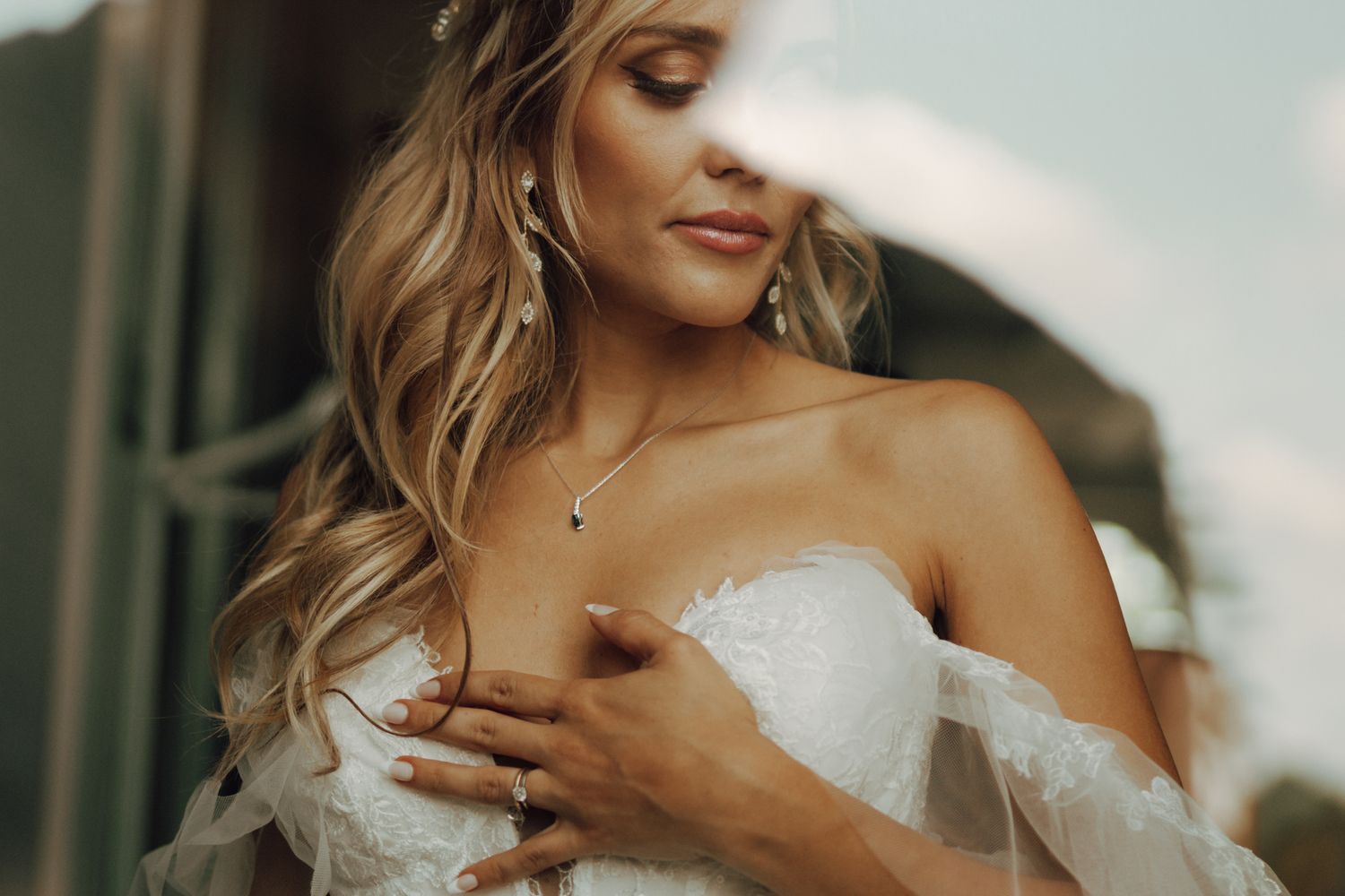 Beautiful blonde bride in strapless lace gown with hand on chest wearing a necklace.