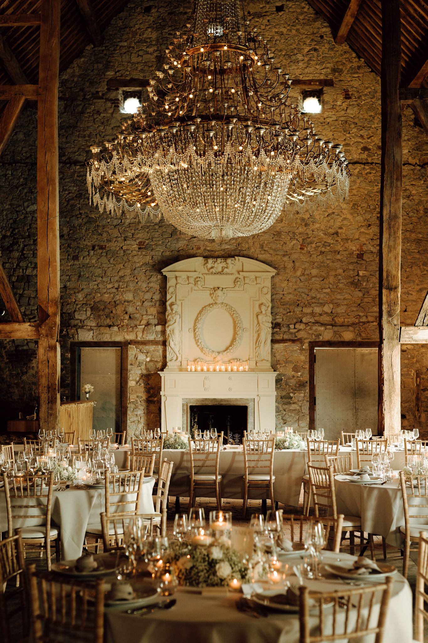 Grand rustic barn wedding reception with crystal chandelier, stone walls, and candlelit round tables.