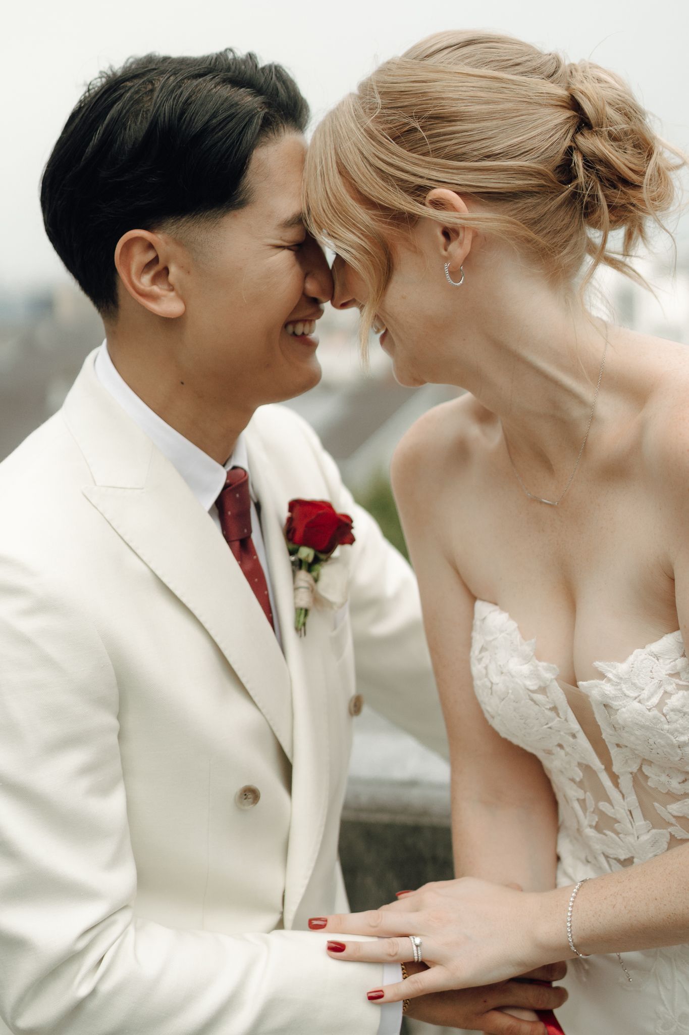 Bride and groom in white suit with red rose boutonniere share romantic moment touching foreheads on wedding day.