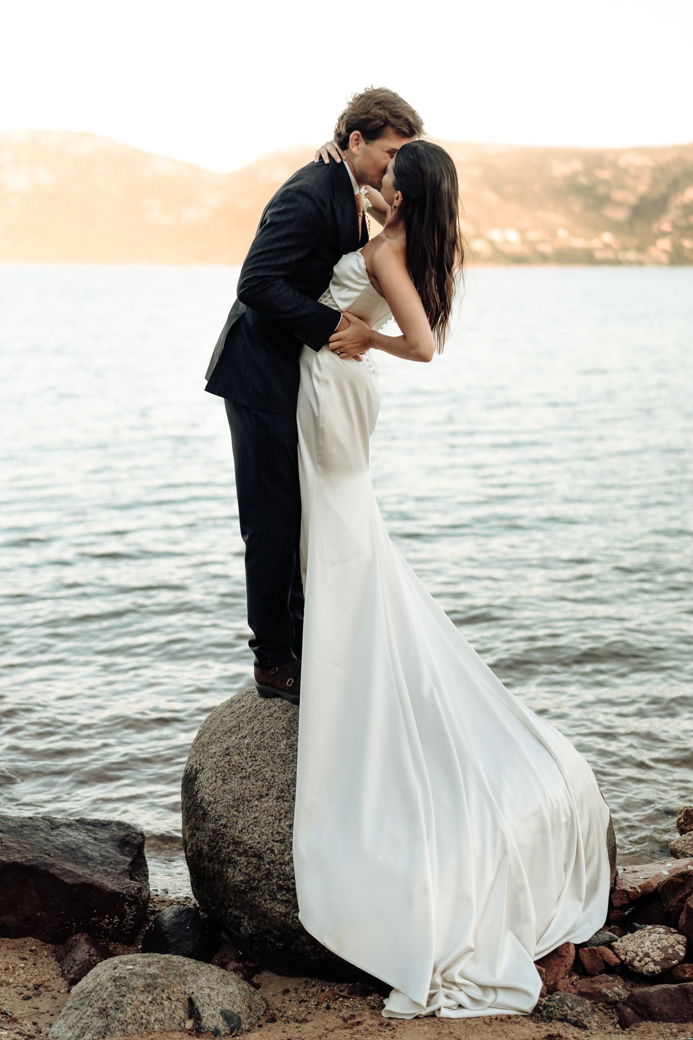 A bride in a white gown and groom in a black suit share a romantic kiss on rocks by a serene lake at sunset.