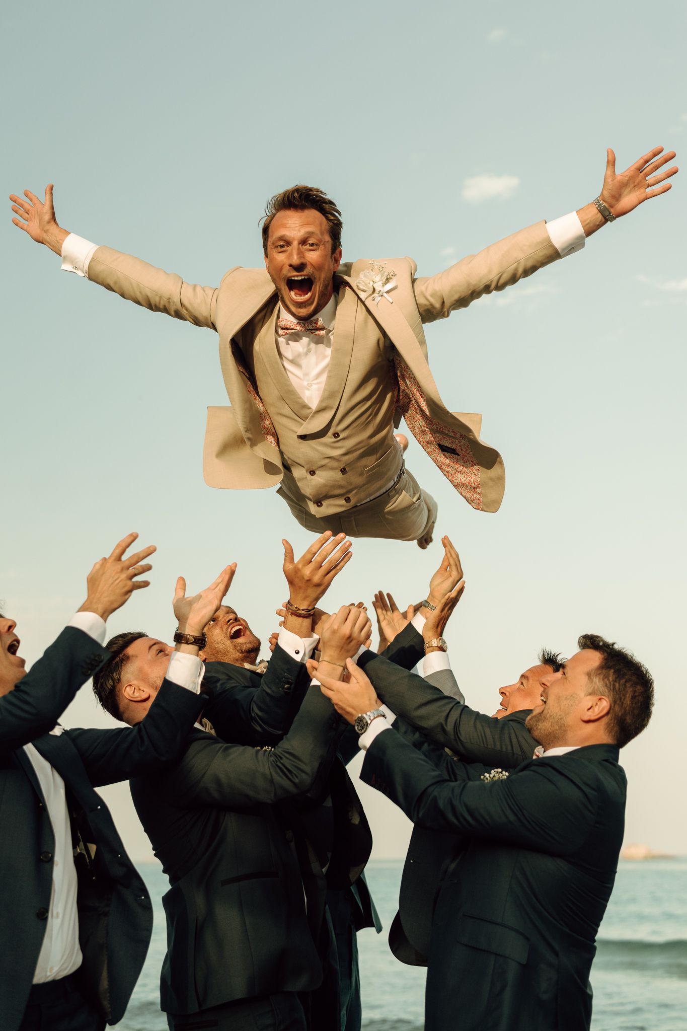 Groomsmen tossing joyful groom in tan suit into the air against a clear blue sky at beach wedding.