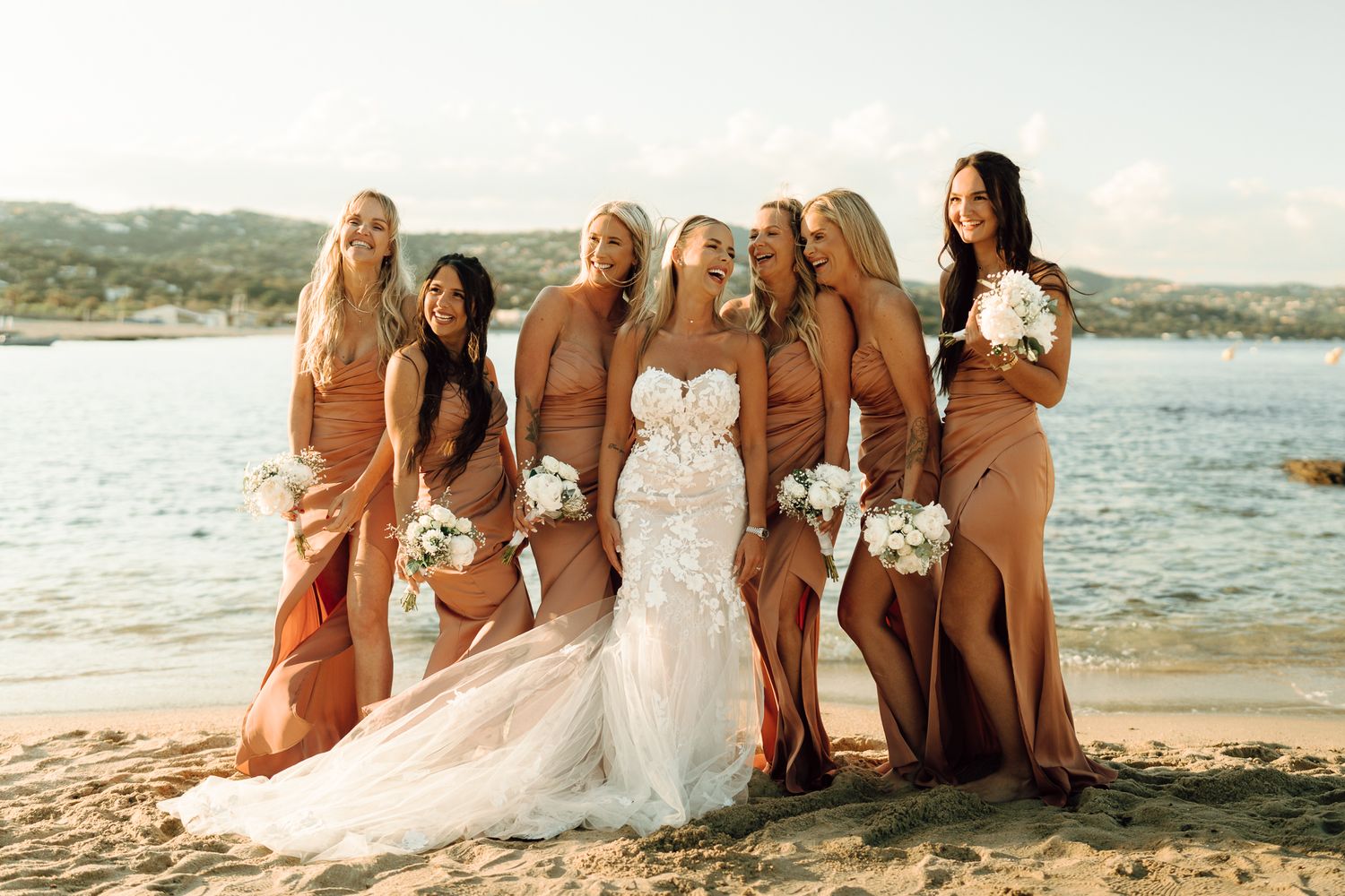 Bride with bridesmaids in rust orange gowns holding white bouquets on a sandy beach at sunset.