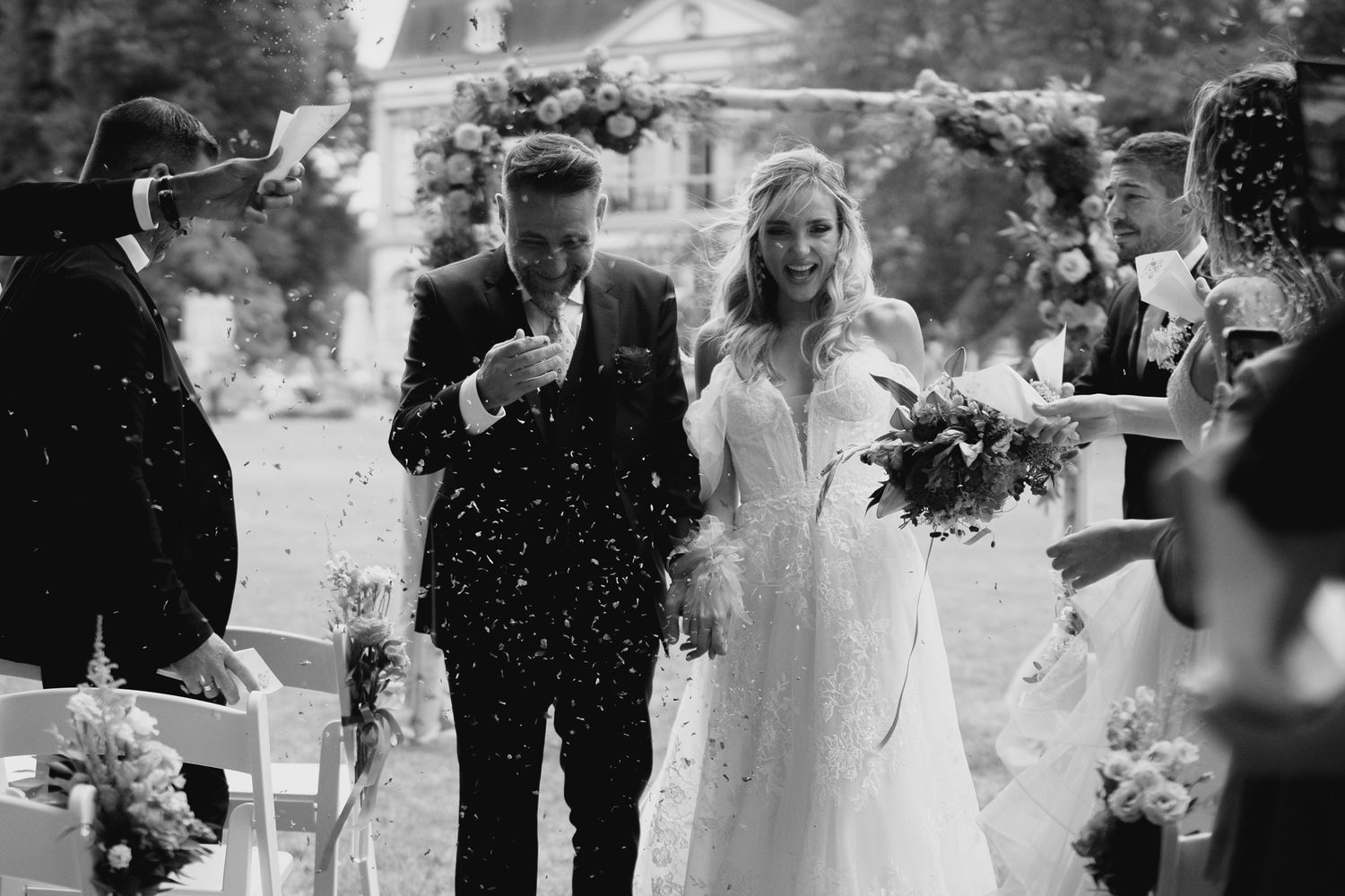 Black and white photo of newlyweds walking down aisle as guests toss confetti at outdoor wedding ceremony.