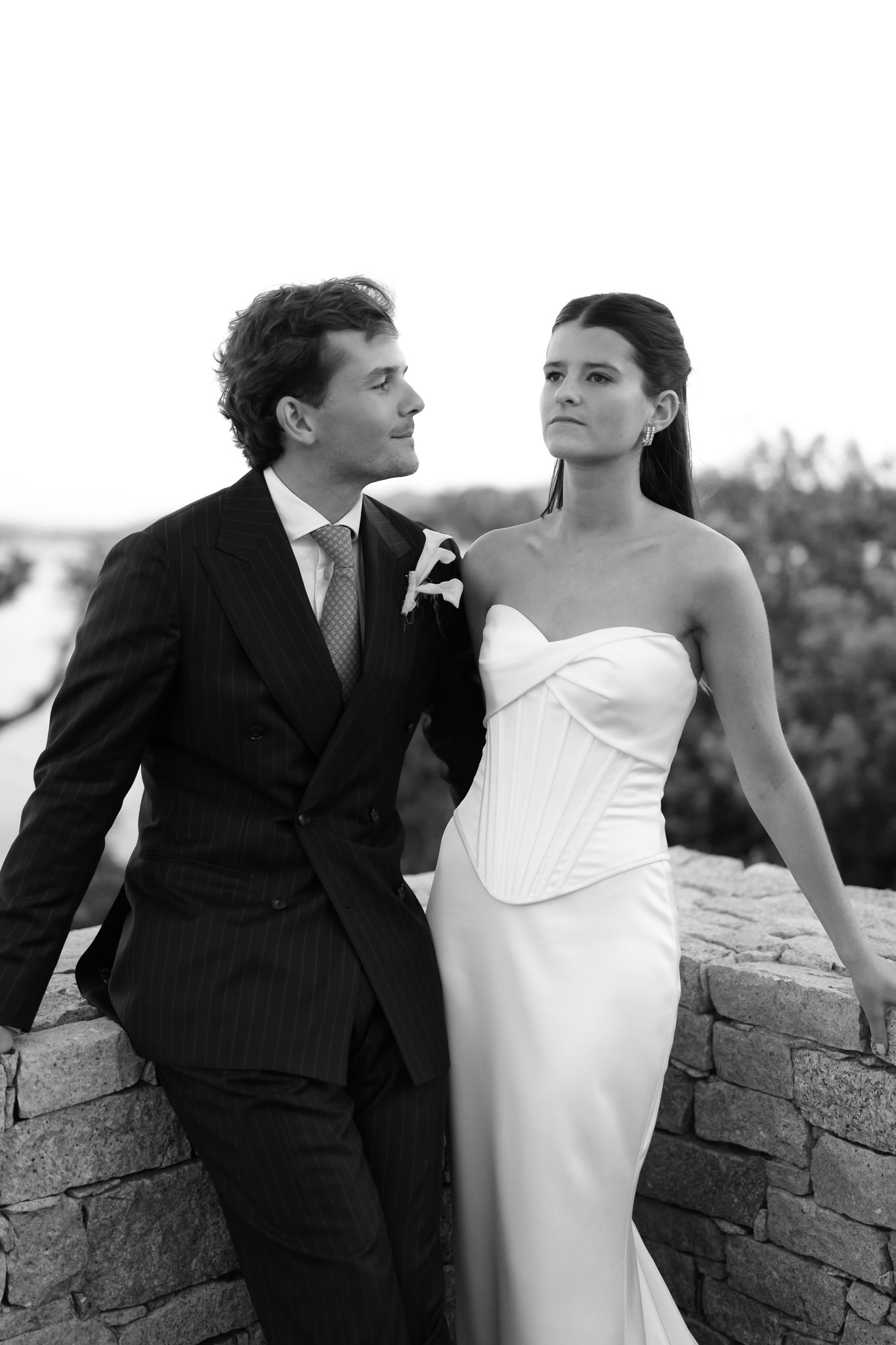 Black and white photo of an elegant bride in strapless gown and groom in suit posing on stone wall outdoors.