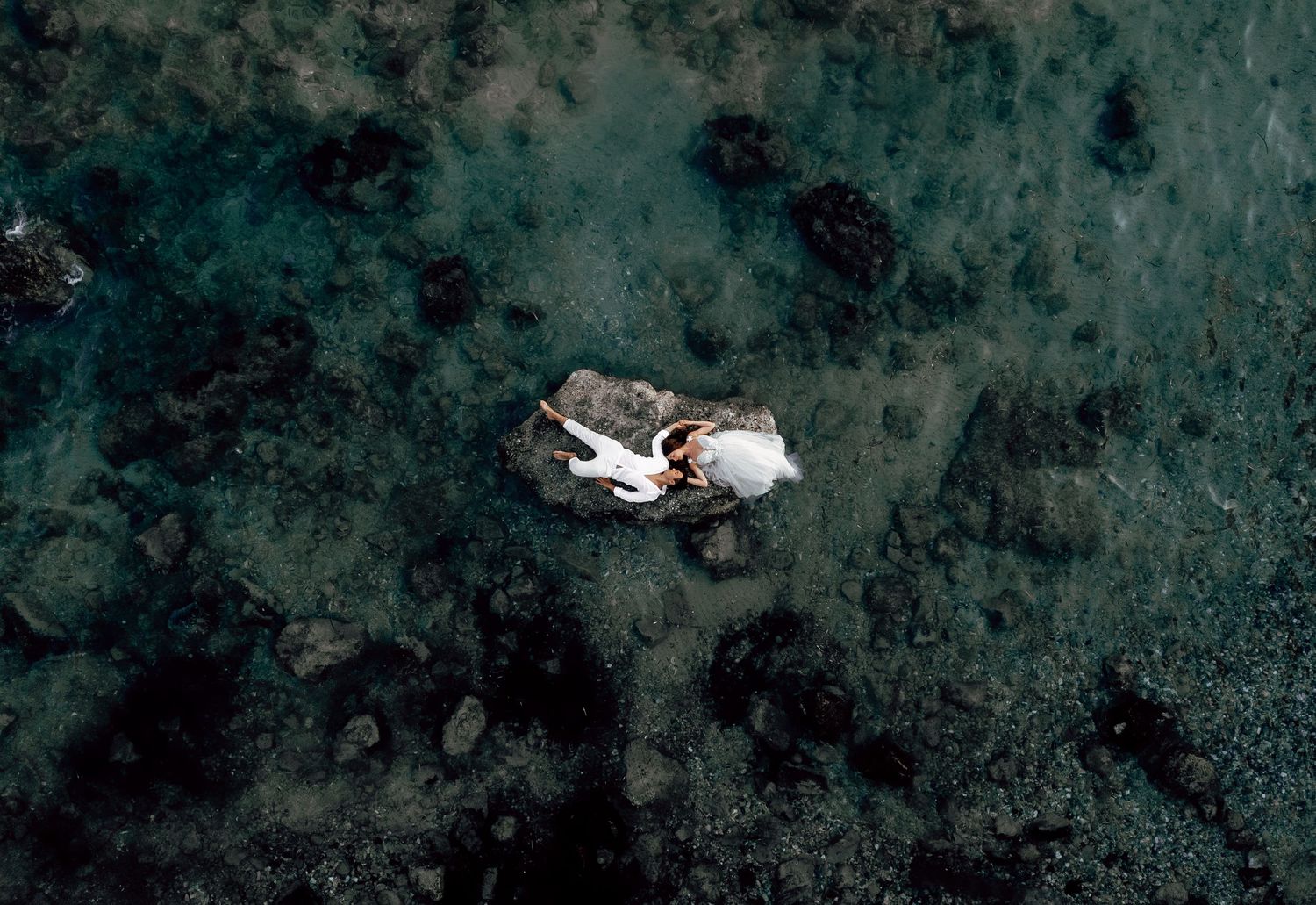 Aerial view of a couple in wedding attire posing on a rocky outcrop surrounded by dark turquoise ocean water.