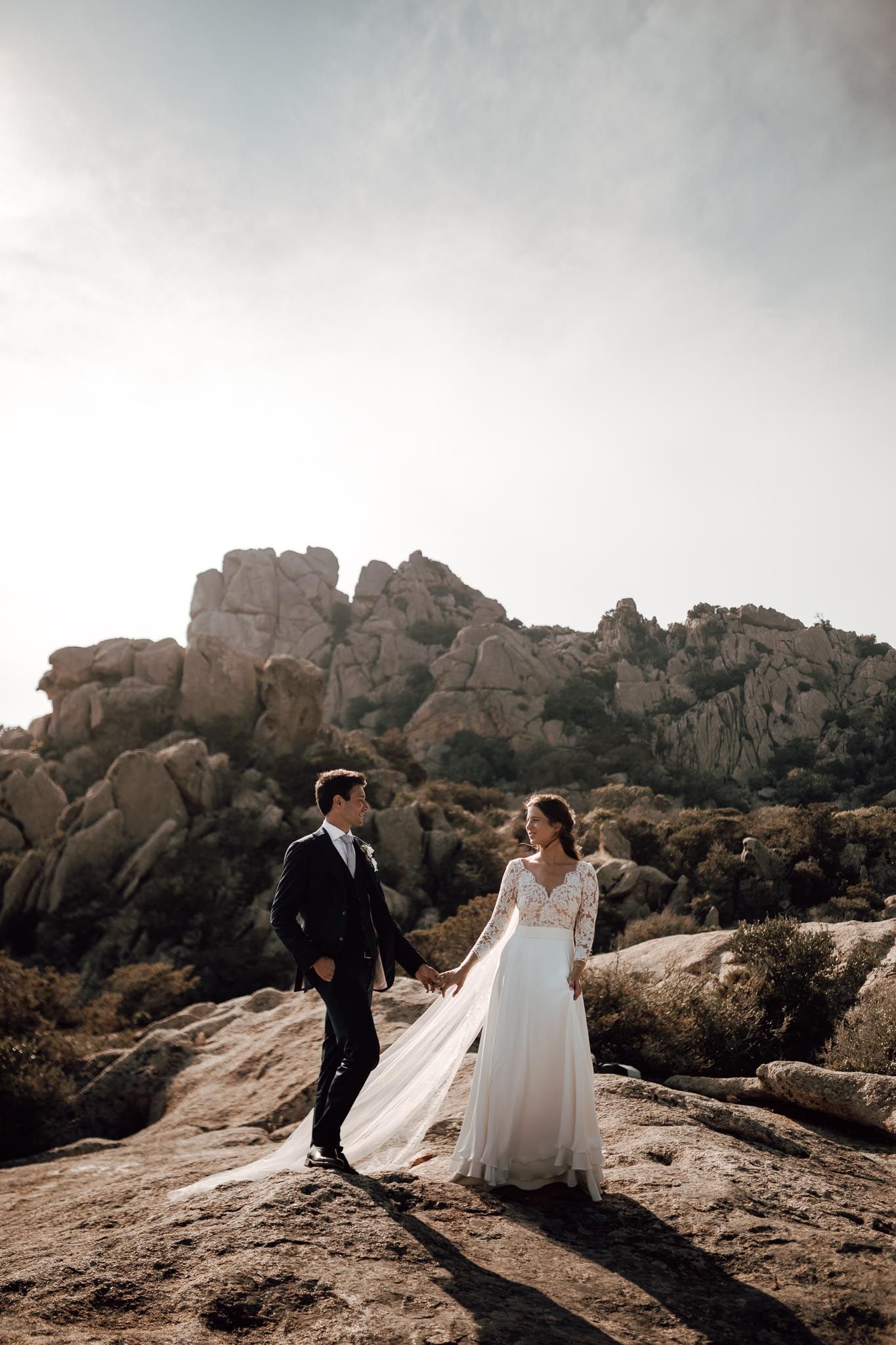 Bride and groom holding hands on rocky landscape with dramatic stone formations backdrop on their wedding day.