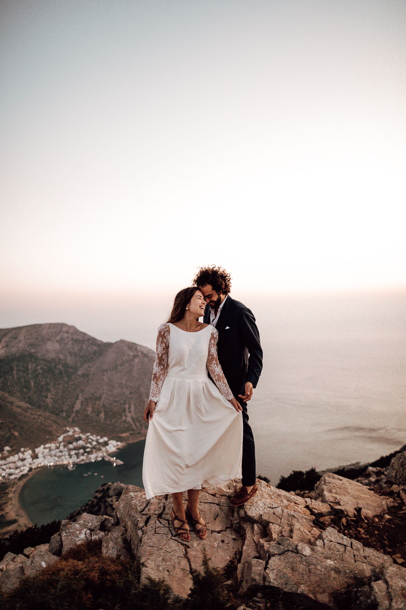 Bride and groom share a romantic moment on a rocky mountaintop with a stunning coastal sunset view.