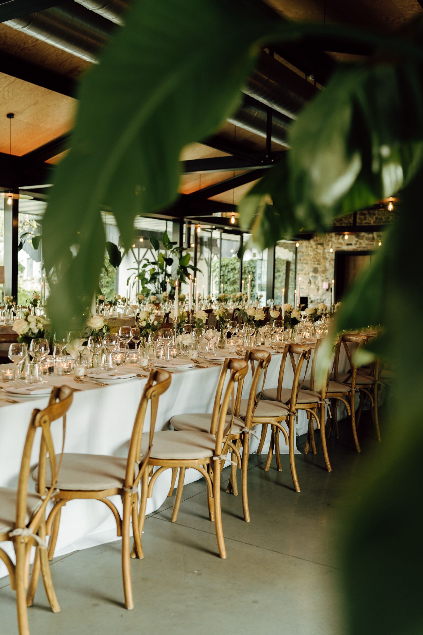 Beautifully set wedding reception tables with wooden cross-back chairs, white florals, and glassware in rustic venue.