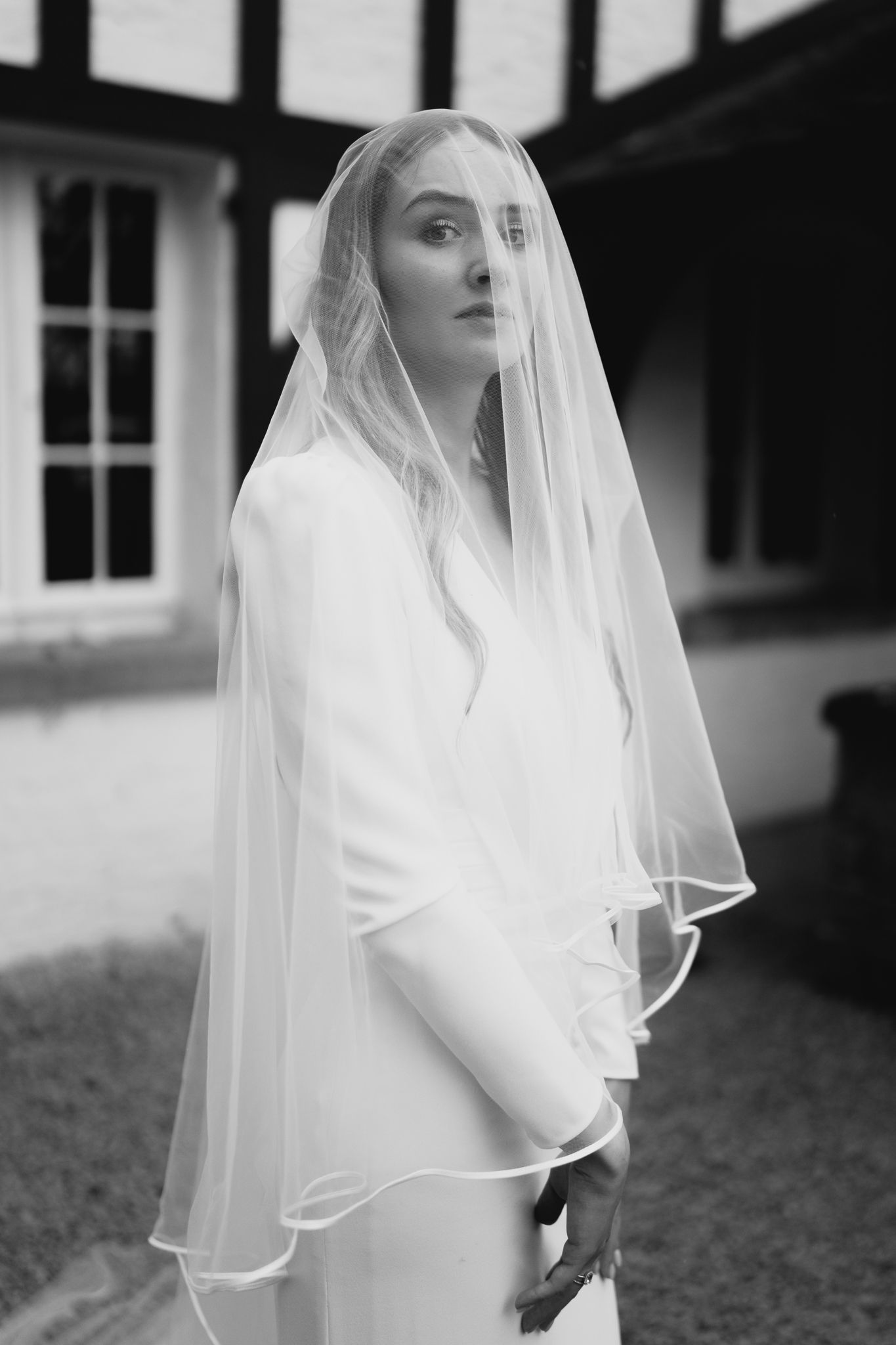 Black and white portrait of a bride wearing a veil over her head outside a half-timbered building.