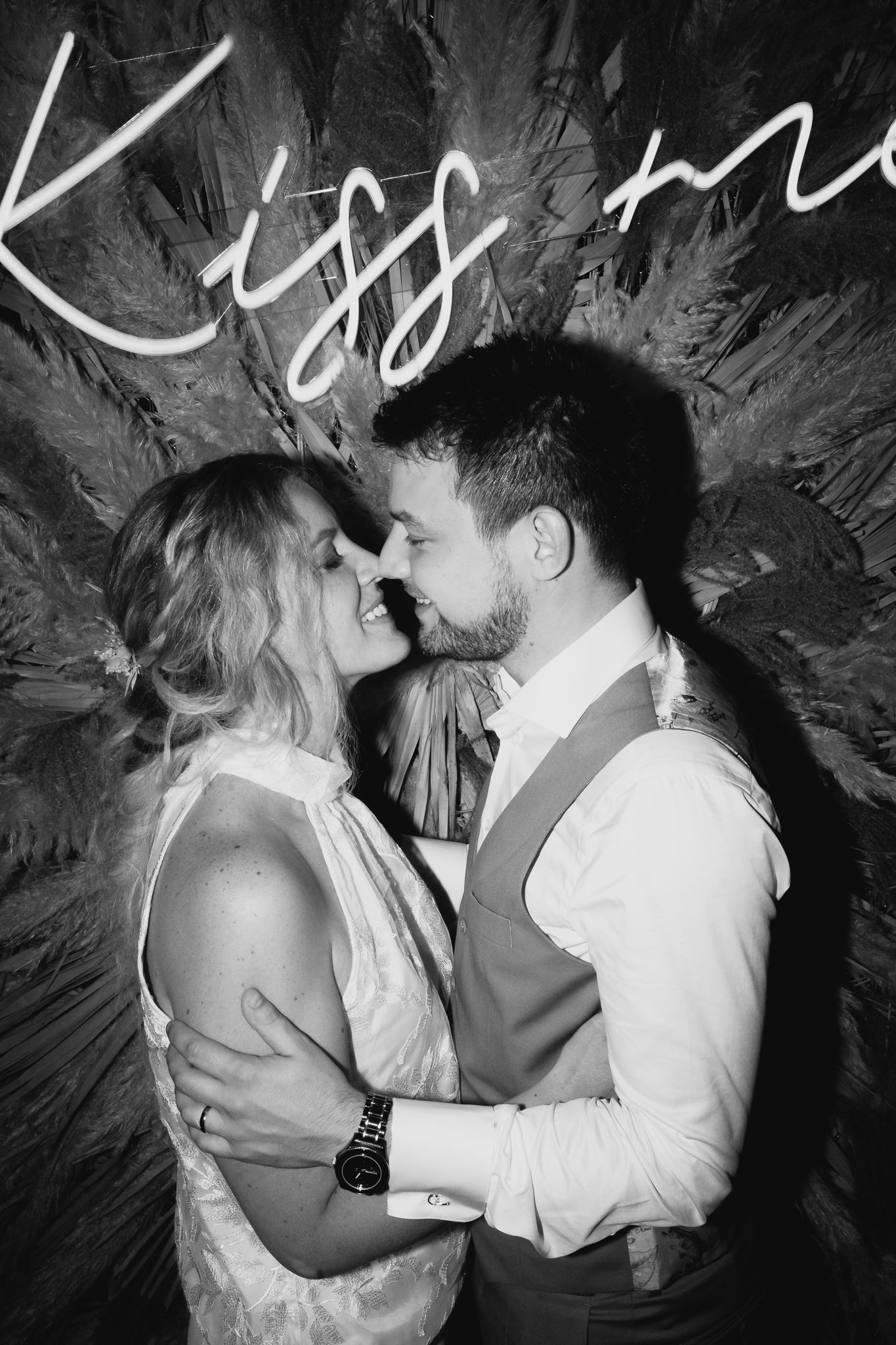 Black and white photo of couple embracing under a Kiss Me neon sign at wedding reception.