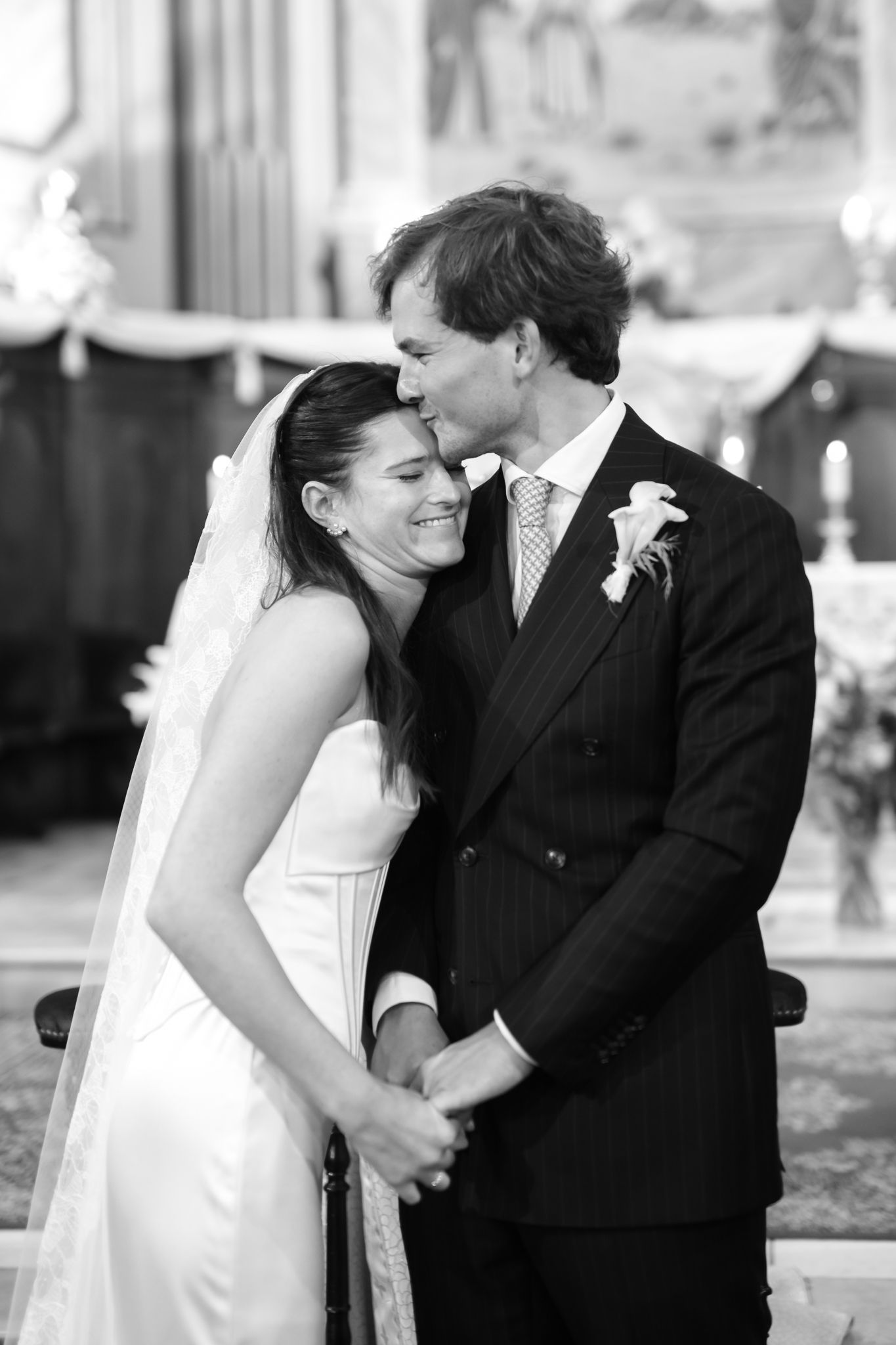 Black and white photo of a groom kissing bride on forehead during wedding ceremony in elegant church.