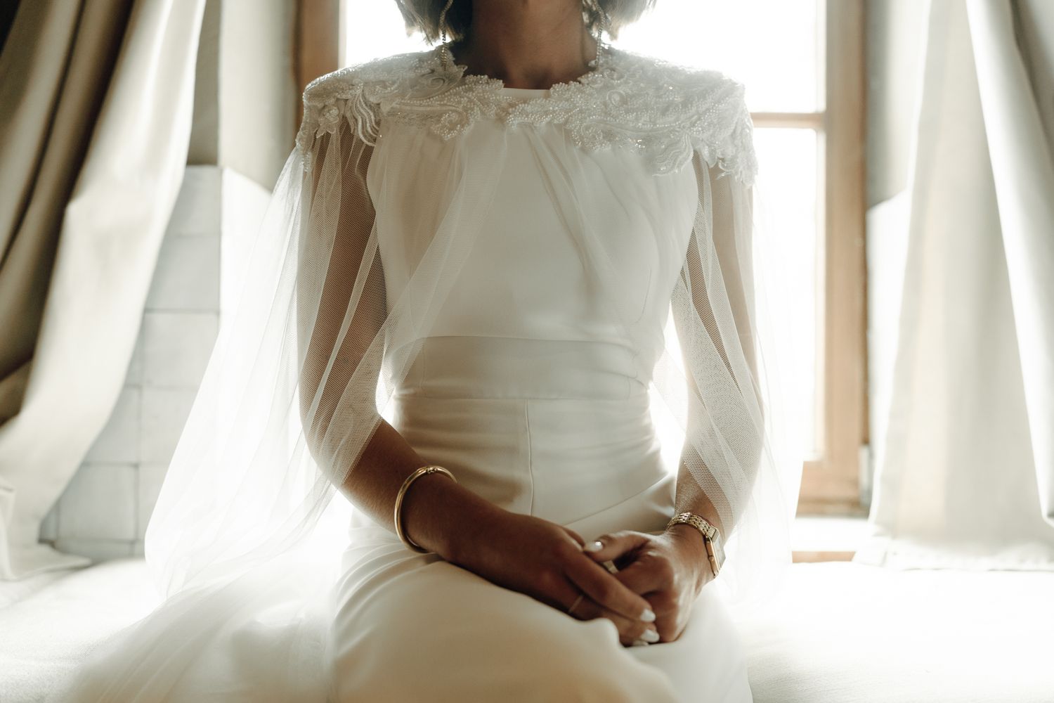 Elegant bride in a white long-sleeve lace wedding gown sitting gracefully near a window on her wedding day.