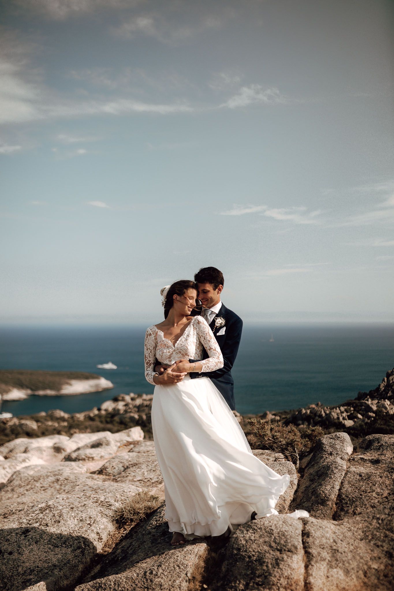 Bride and groom embracing on rocky coastal cliffs with turquoise ocean views during their destination wedding.