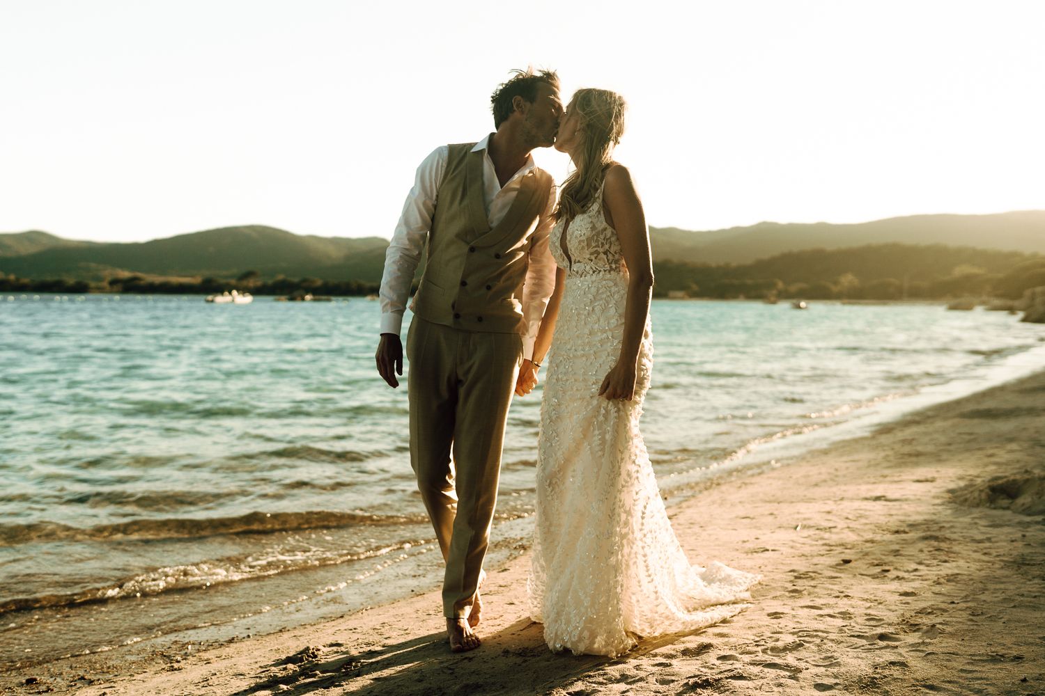 Newlyweds sharing a romantic kiss on a sunlit beach at sunset, with sparkling blue water behind them.