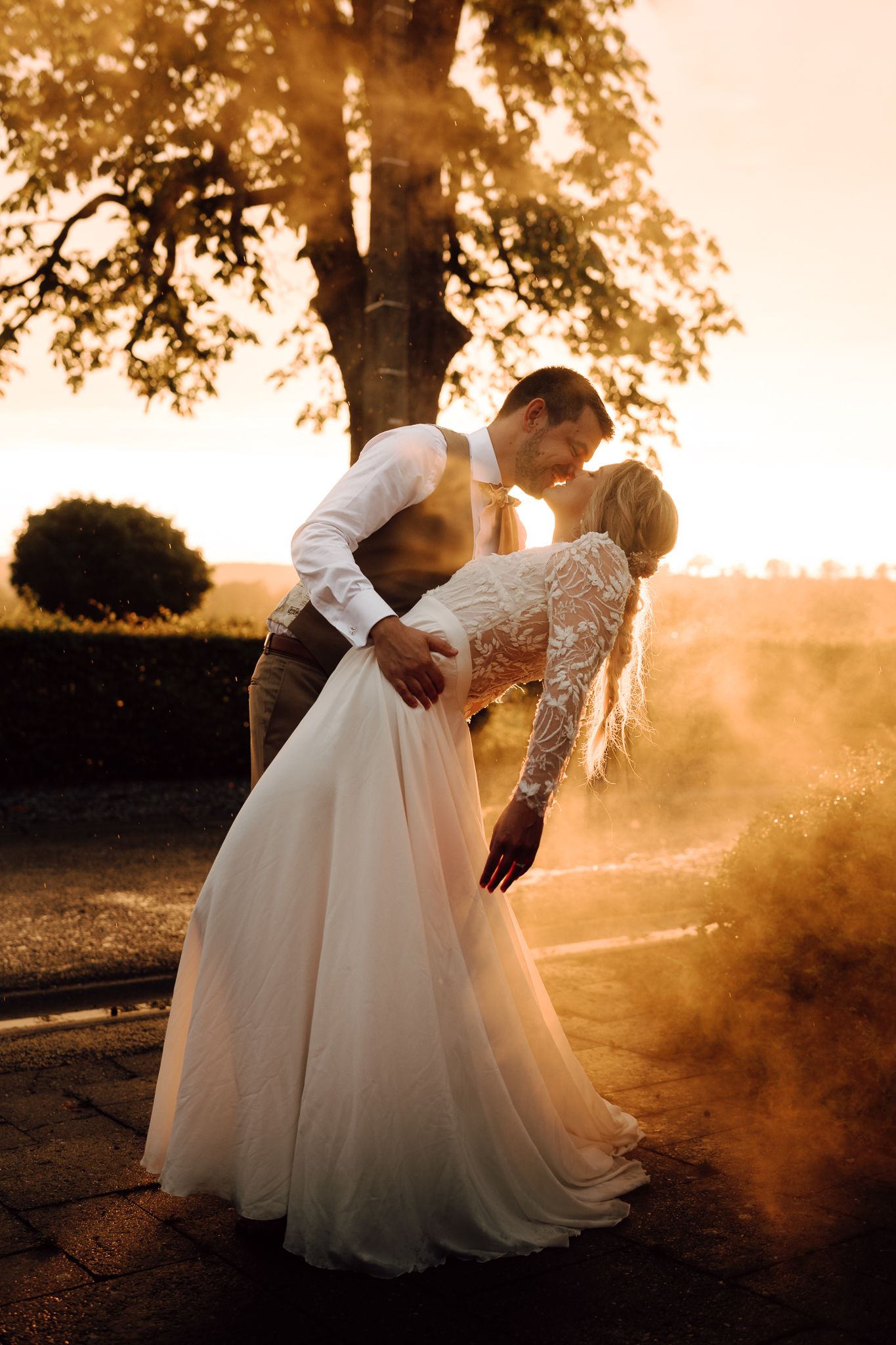 Bride and groom sharing a romantic dip kiss at golden hour sunset, with warm light and mist surrounding them outdoors.