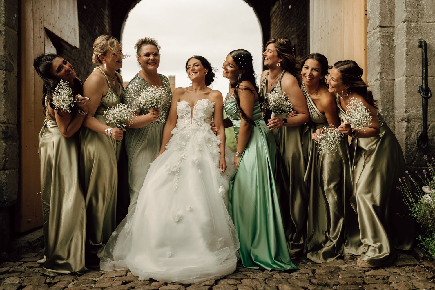 Radiant bride posing with six bridesmaids in sage green satin gowns at a stone archway.
