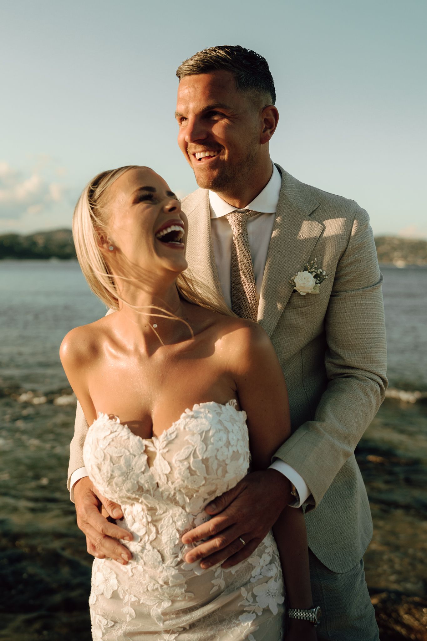 Laughing bride in lace gown embraced by groom in beige suit on rocky shoreline at golden hour.