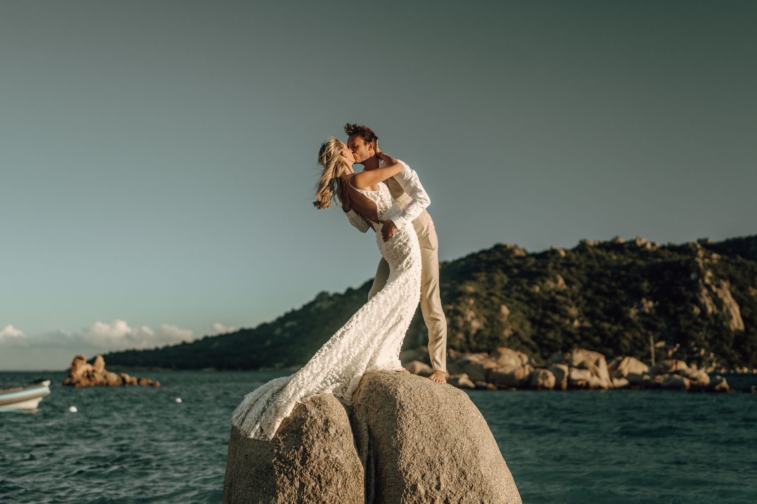 Laughing groom in beige vest holding brides hand near a shimmering sunset ocean on wedding day.