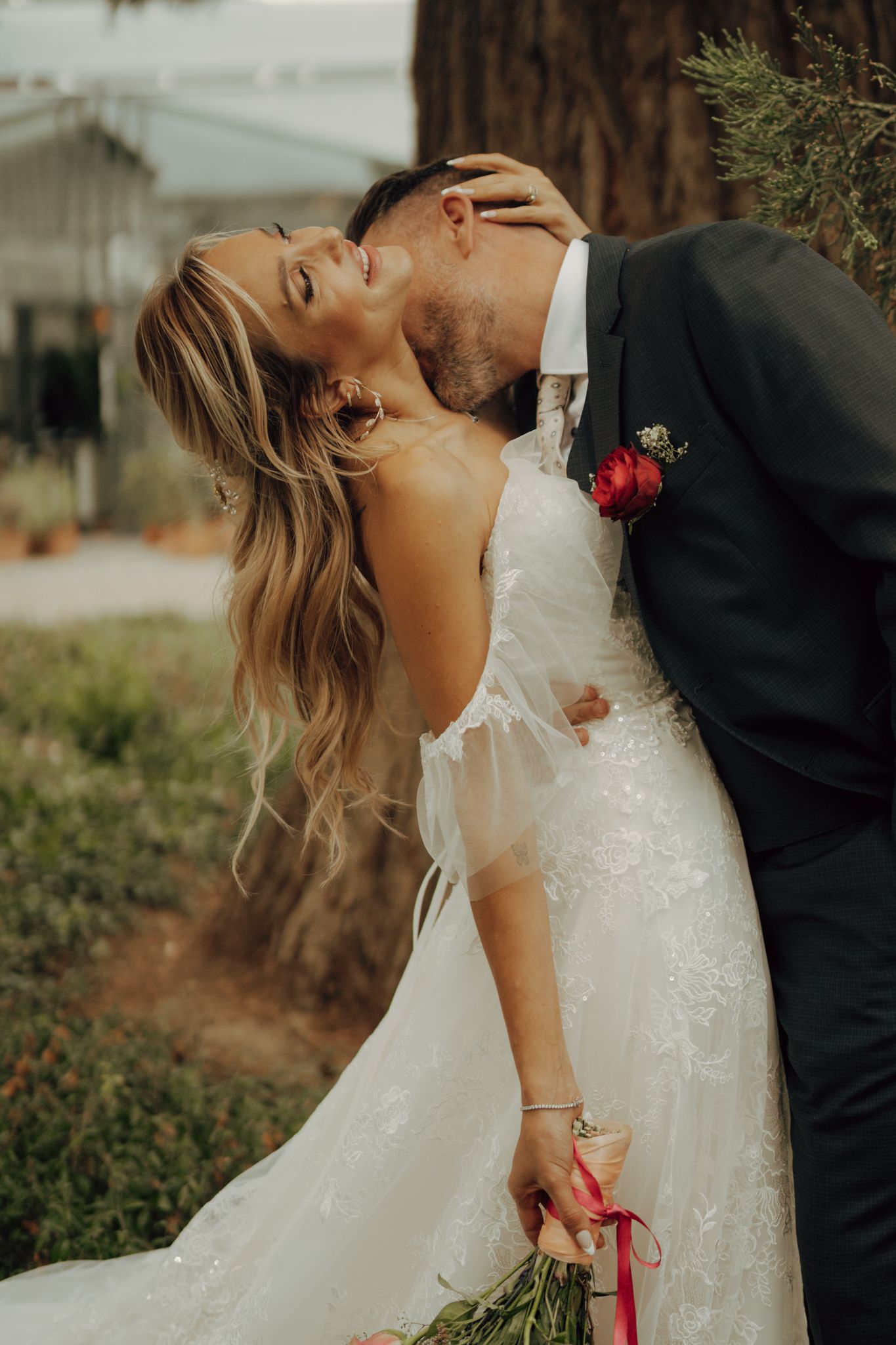 Groom in dark suit kissing bride in white lace gown outdoors near pine tree.