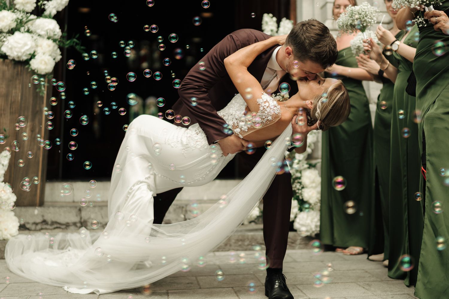 Groom dips bride in romantic kiss surrounded by bubbles and green-dressed bridesmaids outdoors.