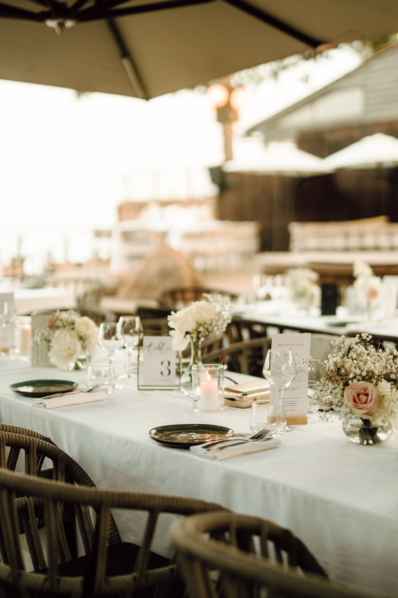 Elegant outdoor wedding reception table set with white linens, floral centerpieces, and glassware under umbrellas.