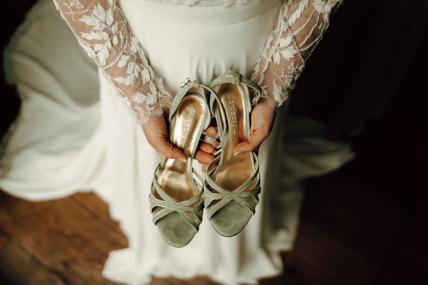 Bride in lace wedding dress holding sage green strappy heels in her hands.