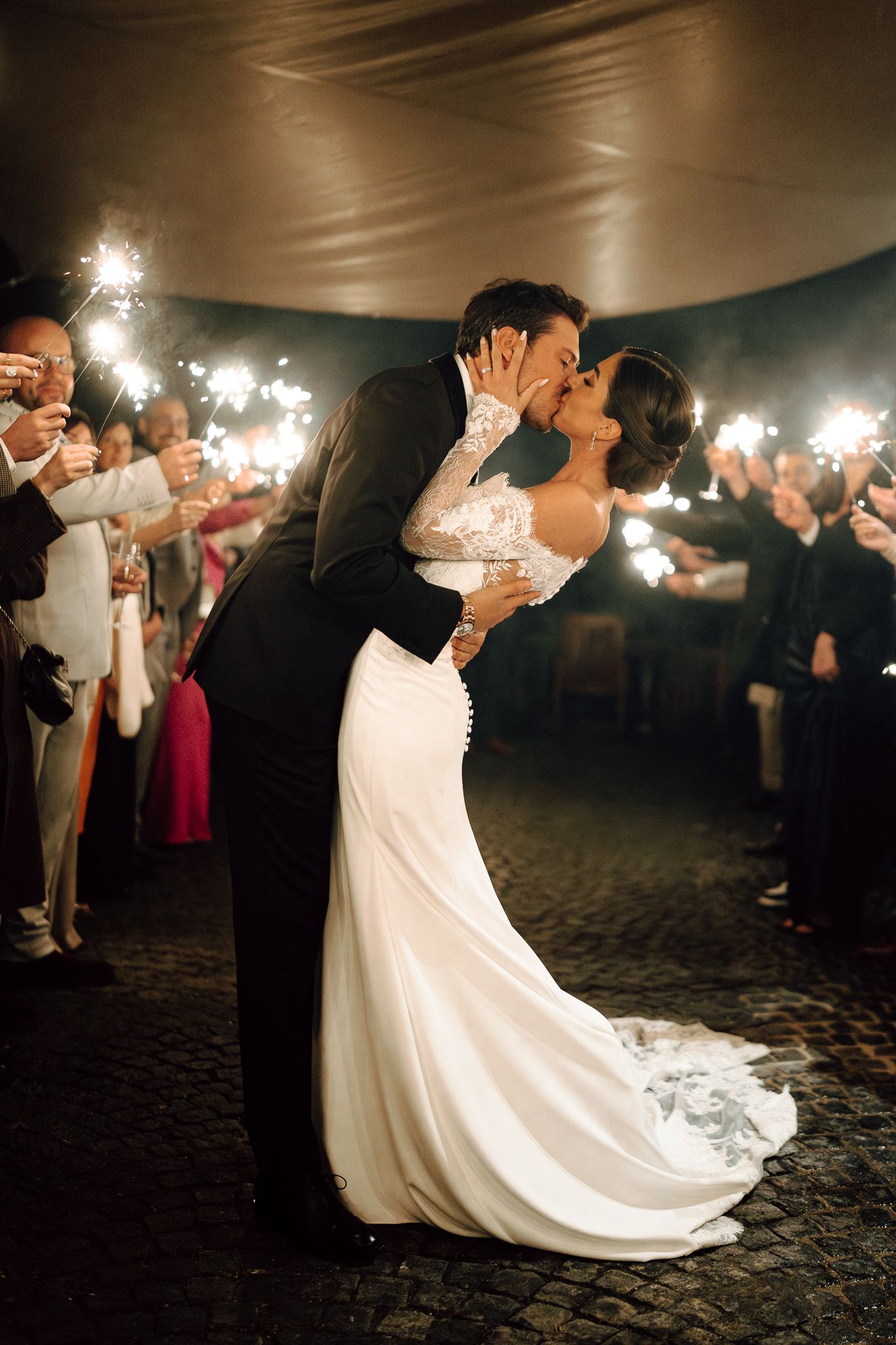 Bride and groom sharing passionate kiss surrounded by guests holding sparklers at night reception.