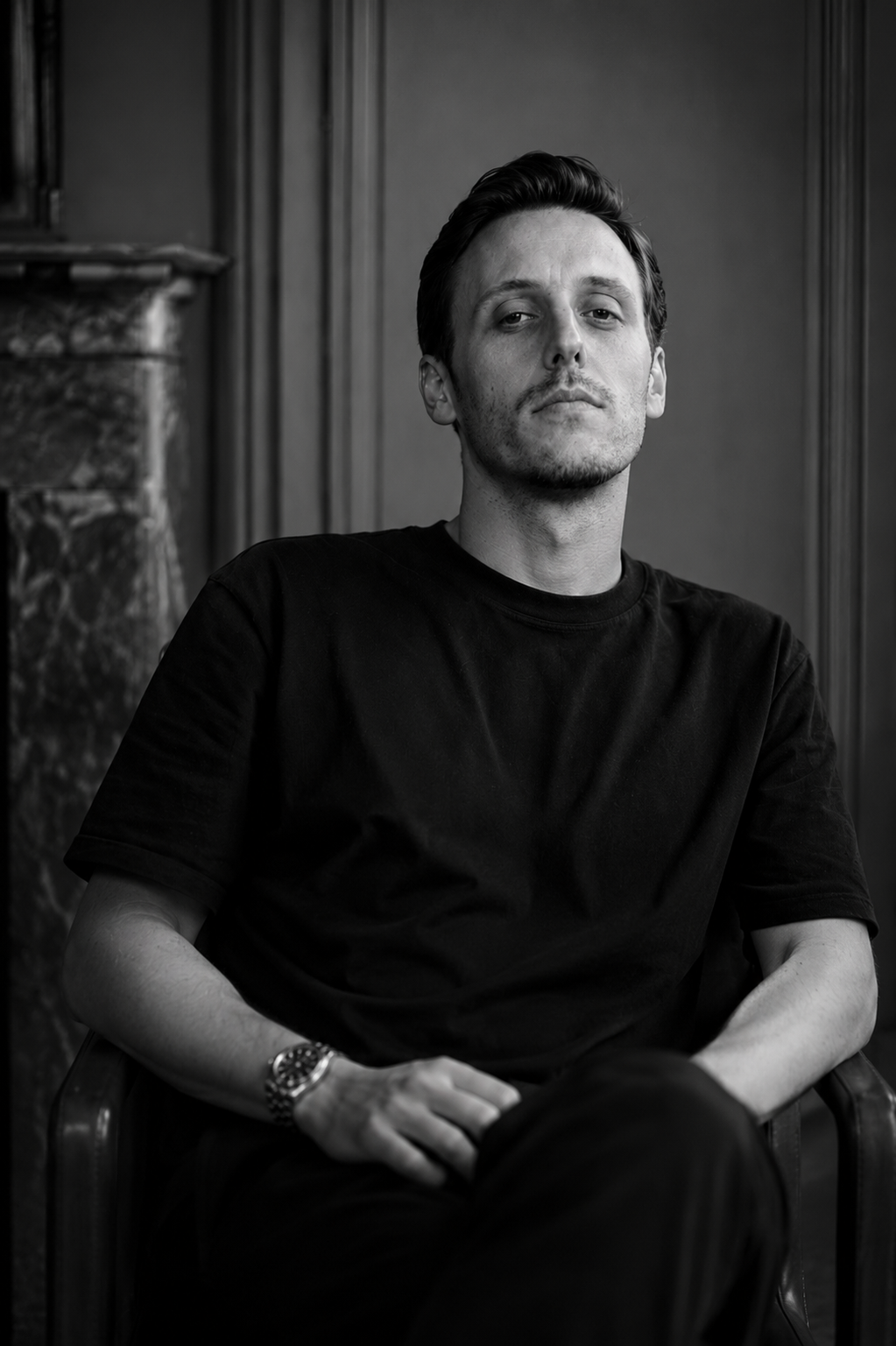 Black and white portrait of a young man in a black t-shirt seated in a leather chair wearing a watch.