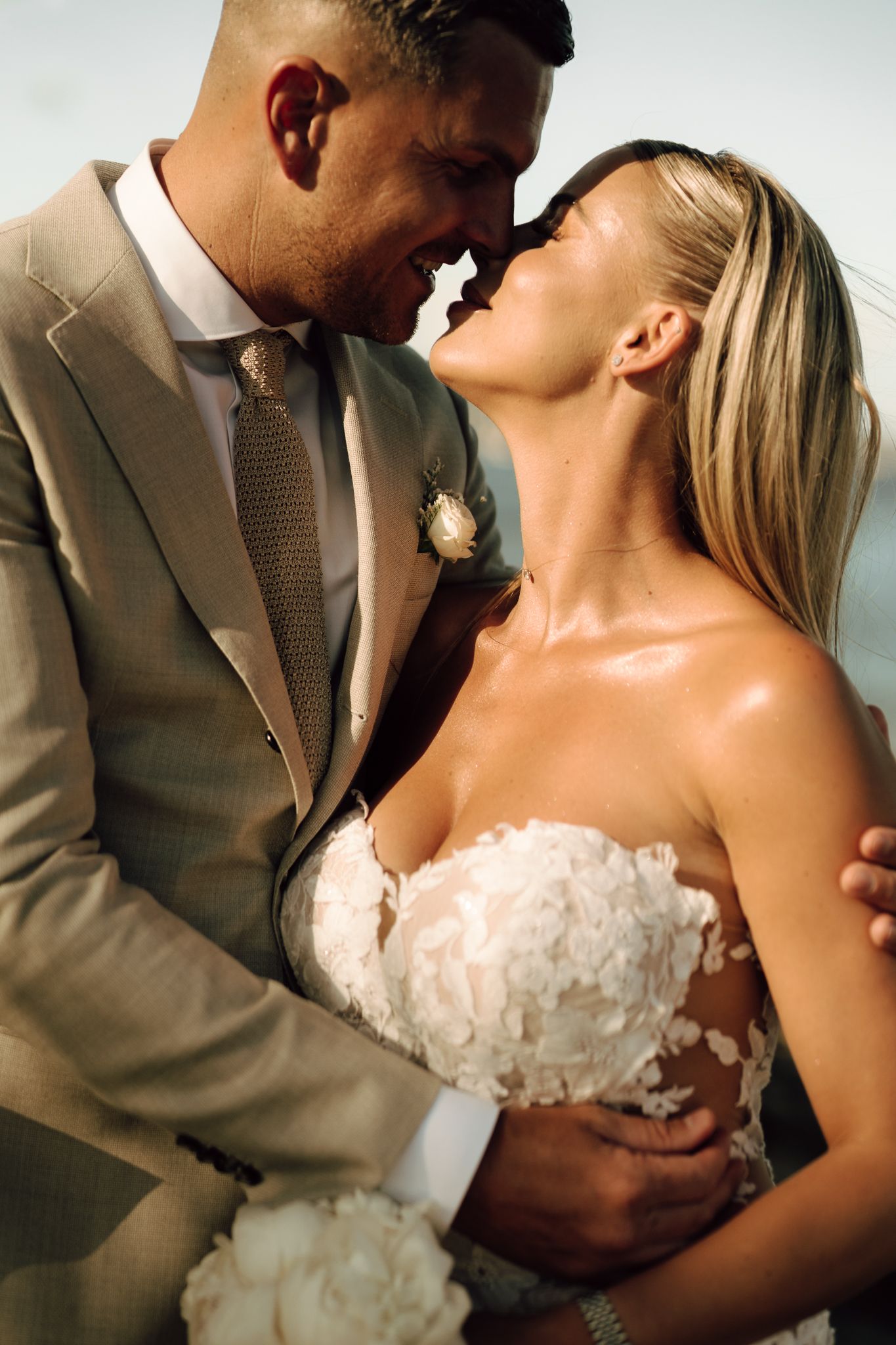Bride in floral strapless gown and groom in tan suit share a romantic close moment outdoors.
