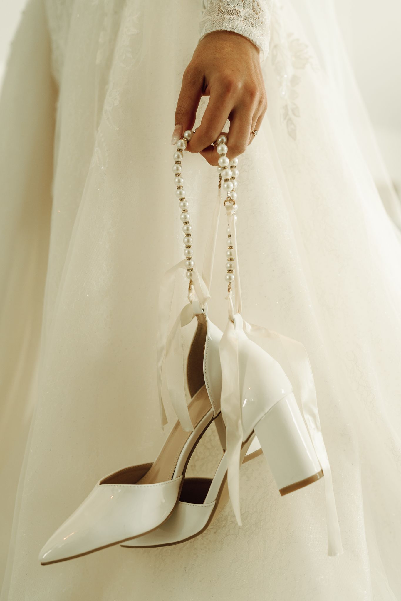 Bride in lace wedding gown holding white pointed-toe heels adorned with pearl and ribbon details.