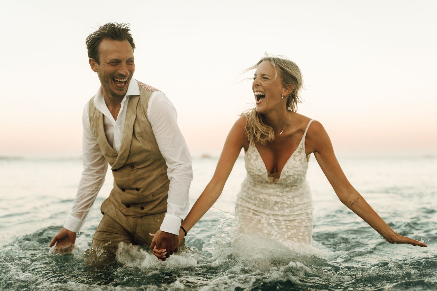 Laughing newlyweds in wedding attire splash joyfully in ocean waves at sunset beach wedding.
