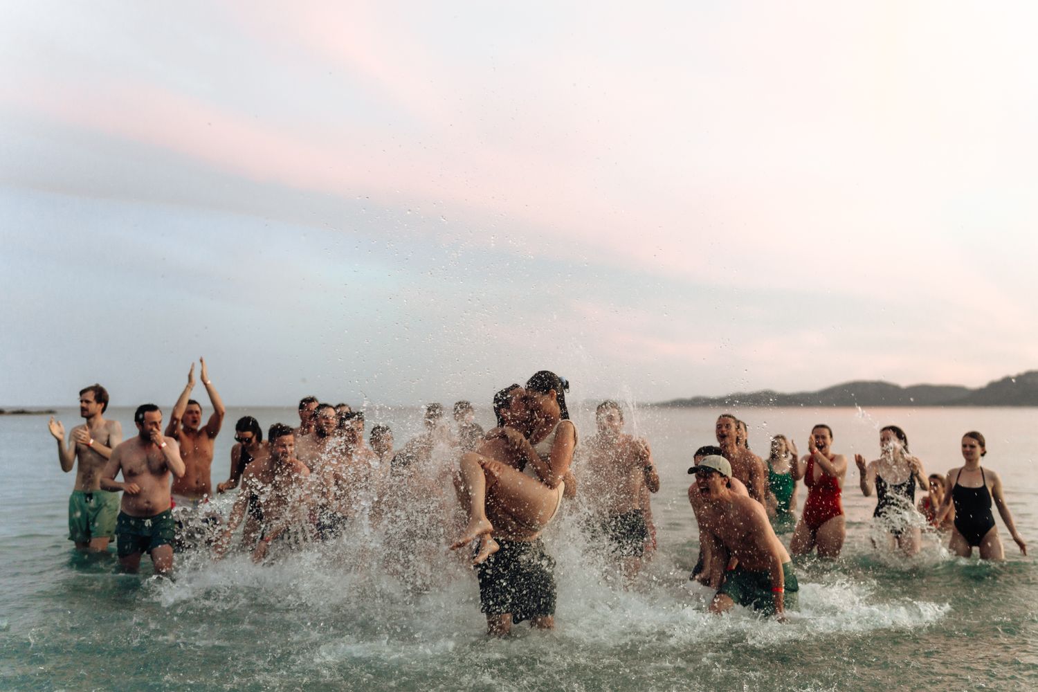 Group of joyful people splashing in ocean waves at sunset during a beach celebration.