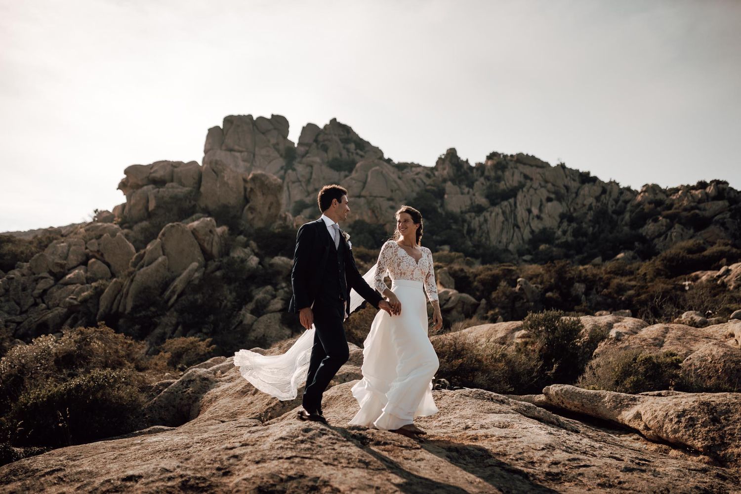 Wedding couple walking hand in hand across rocky terrain with stunning boulder formations in the background.