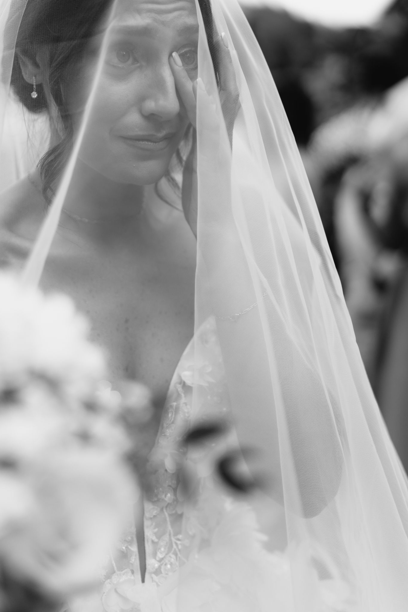 Emotional black and white portrait of tearful bride peeking through flowing wedding veil.